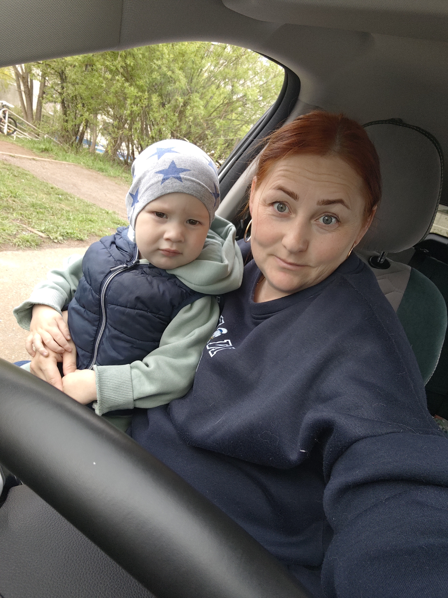 A woman and a young child are seated inside a vehicle, possibly in the driver's seat. The woman has red hair and wears a dark blue sweatshirt and hoop earrings. The child, wearing a blue vest over a green sweatshirt and a gray hat with blue stars, is in her lap, their hands together. The background shows a glimpse of a road, grass, trees with green leaves, and possibly a building or structure. The location is Markovskii, Russia. The steering wheel is visible in the foreground.