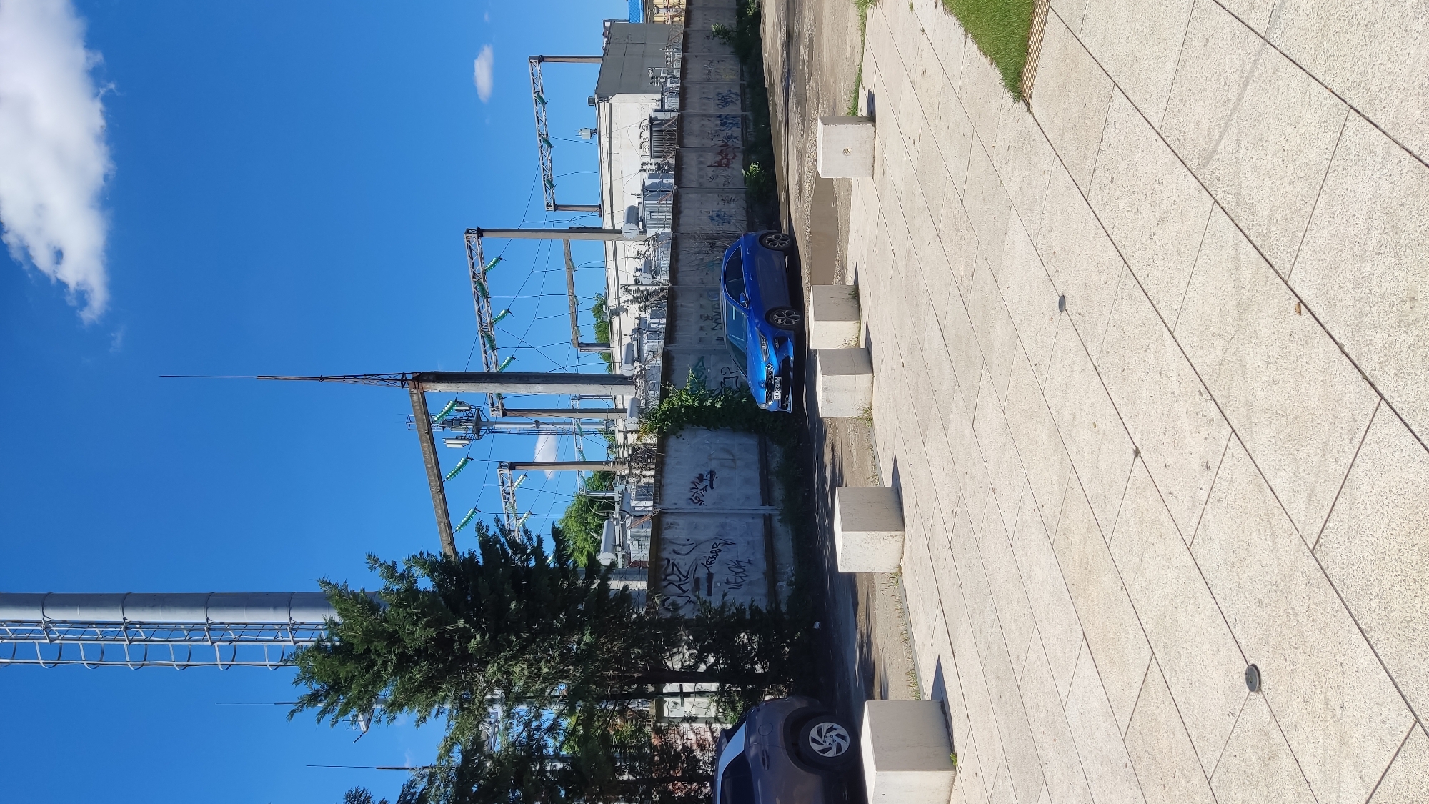The image depicts an outdoor urban or industrial scene in Krasnodar, Russia, under a clear blue sky. The main subjects are an electrical substation, parked cars, and a pedestrian pathway.
In the background, a large electrical substation is visible, characterized by numerous metal towers, power lines, and insulators. Concrete buildings are integrated into the substation complex. A tall, slender lightning rod extends upwards on the left. Separating the substation from the foreground is a concrete wall, extensively covered in graffiti. Partially decipherable text on the wall includes "KREOR" and "VECH."
Two cars are parked parallel to this wall. A bright blue hatchback car is prominently featured in the mid-ground, facing right. Its license plate, "50-70 BB 93," is clearly visible, with "93" indicating the Krasnodar Krai region. A dark grey or black car is partially visible in the bottom right corner of the frame.
The foreground consists of a wide pedestrian pathway paved with light-colored concrete tiles, which appear to be in good condition. Several rectangular concrete bollards are spaced along the edge of the path, likely serving as barriers. Green trees and bushes are scattered throughout the scene, providing some natural elements amidst the industrial setting.
The time of day is clearly daytime, likely morning or afternoon, given the bright illumination and strong shadows. The weather is sunny with a clear blue sky and a few fluffy white clouds. No human activity or specific event is taking place; the scene is static.