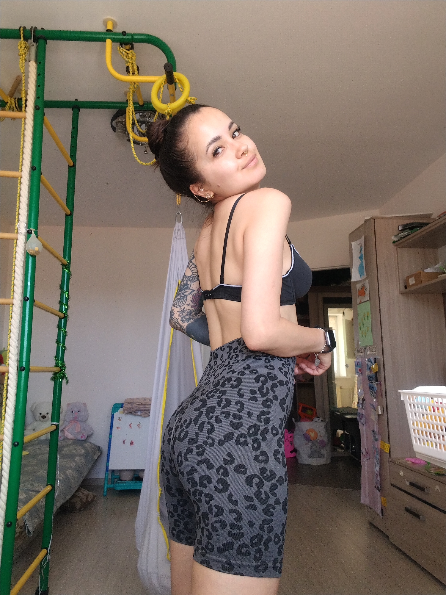 This media file captures a young woman with dark hair styled in a bun, wearing a black sports bra and grey leopard-print shorts. She is posing in an indoor setting, looking over her shoulder towards the camera with a slight smile. Her left arm is adorned with tattoos.
In the background, a green and yellow indoor playground structure with ropes and rings is visible on the left. To the right, a wardrobe with various items on its shelves and doors stands against a wall. A white laundry basket is placed on top of a dresser. Two stuffed animals, a white bear and a pink bunny, sit on a gray mat in the left background. A small whiteboard with colorful drawings is also present. The lighting suggests it is daytime. The overall scene appears to be a room in a home, likely a child's bedroom or a multi-purpose recreational space.