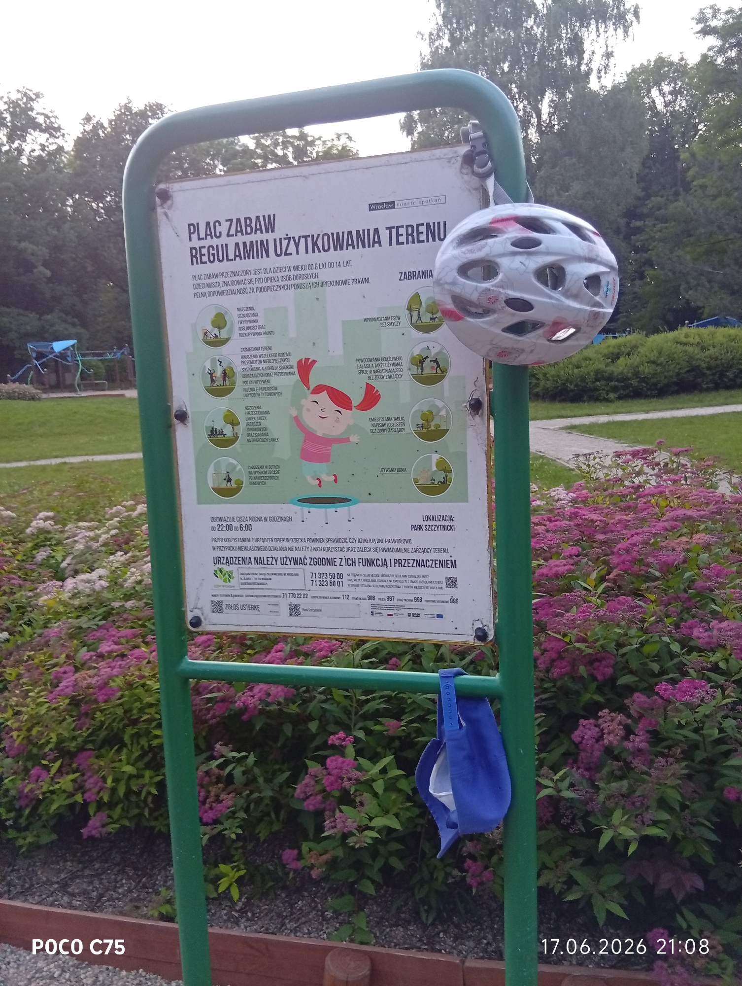 The image shows a park in Wrocław, Poland, featuring a sign that details the regulations for a playground. The sign, mounted on a green metal frame, is partially obscured by a white bicycle helmet hanging from the frame. The helmet is white with red and pink floral designs. The sign itself contains text in Polish and illustrations depicting prohibited activities on the playground, such as climbing on the outside of structures or using equipment improperly. It also specifies that children under the age of 6 must be supervised by an adult. The playground area, indicated by the sign, is surrounded by lush greenery, including a bed of vibrant pink flowers in the foreground. The time stamp "17.06.2026 21:08" is visible in the bottom right corner, suggesting a late evening setting. A blue cloth mask is also hanging from the green frame below the helmet.