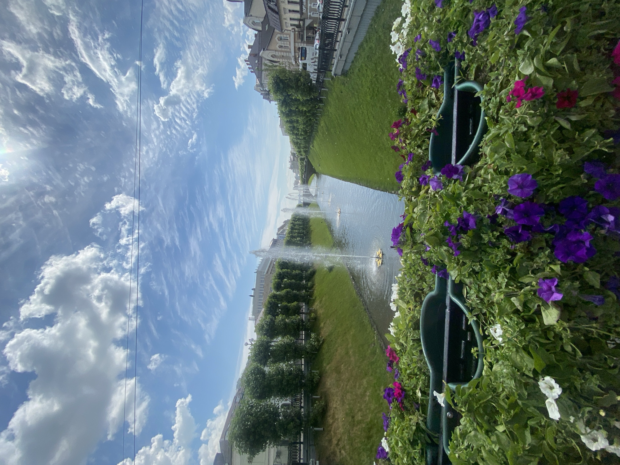 The image depicts a canal in Kazan, Russia, with several fountains spraying water. The canal is lined with green grass and a row of neatly trimmed trees. In the foreground, there is a planter filled with colorful flowers, including purple, pink, and white petunias. Beyond the trees, there are buildings with traditional architecture, suggesting a historic part of the city. The sky is blue with scattered white clouds, and the sun is shining, casting a bright light on the scene. A few vehicles are visible on the street alongside the canal.