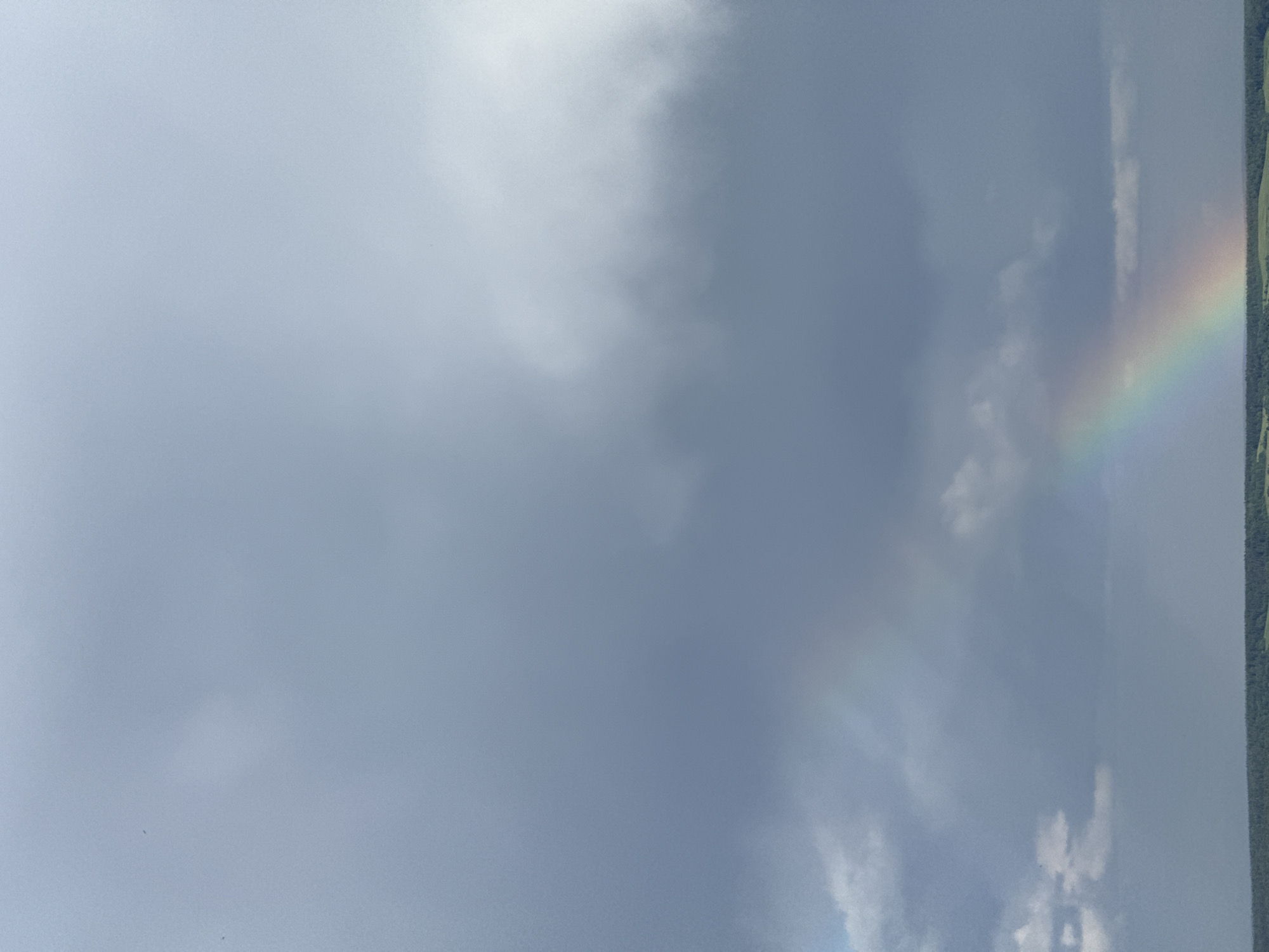 The image depicts a cloudy sky with a rainbow appearing on the right side of the frame. The rainbow's colors are visible, with the arc ending near a line of trees or vegetation. The sky is predominantly gray with varying cloud formations, including brighter patches and denser, darker areas.