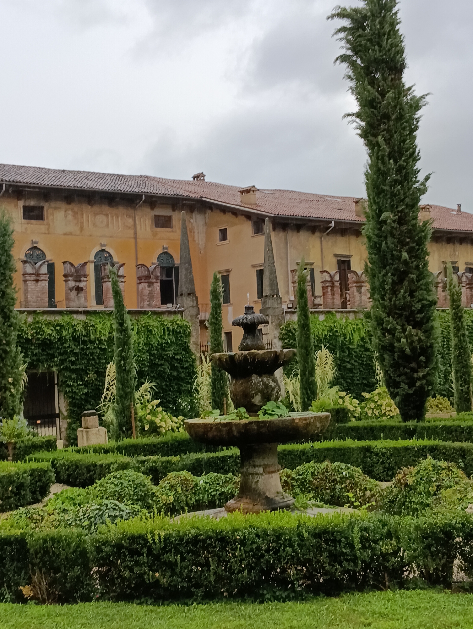 The image depicts a view of the Giardino Giusti in Verona, Italy. The scene features a formal Italian garden with meticulously manicured hedges, topiaries, and a stone fountain in the foreground. Behind the garden, a section of the historic Palazzo Giusti is visible. The palace facade is painted in warm, earthy tones and features arched windows and decorative architectural elements. Lush greenery, including tall cypress trees and ivy-covered walls, cascades down the sides of the building and frames the garden. The sky is overcast, suggesting a cloudy day. There are no people or animals present in the image. The overall impression is one of serene beauty and historical grandeur.