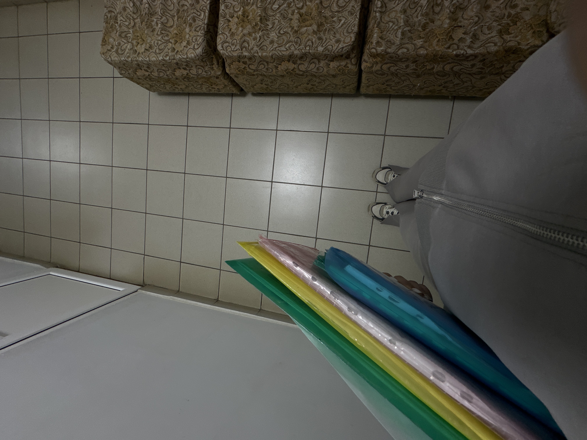 The image presents a top-down view. The scene appears to be indoors, with a tiled floor, possibly in a public building in Mozhga, Russia. Three floral pattern upholstered blocks are visible in the upper left corner. A person wearing light grey clothing and sneakers stands on the tiled floor. The person is holding a stack of colored plastic folders (green, yellow, pink, blue) over what appears to be a white surface. The context suggests an office or institutional environment.