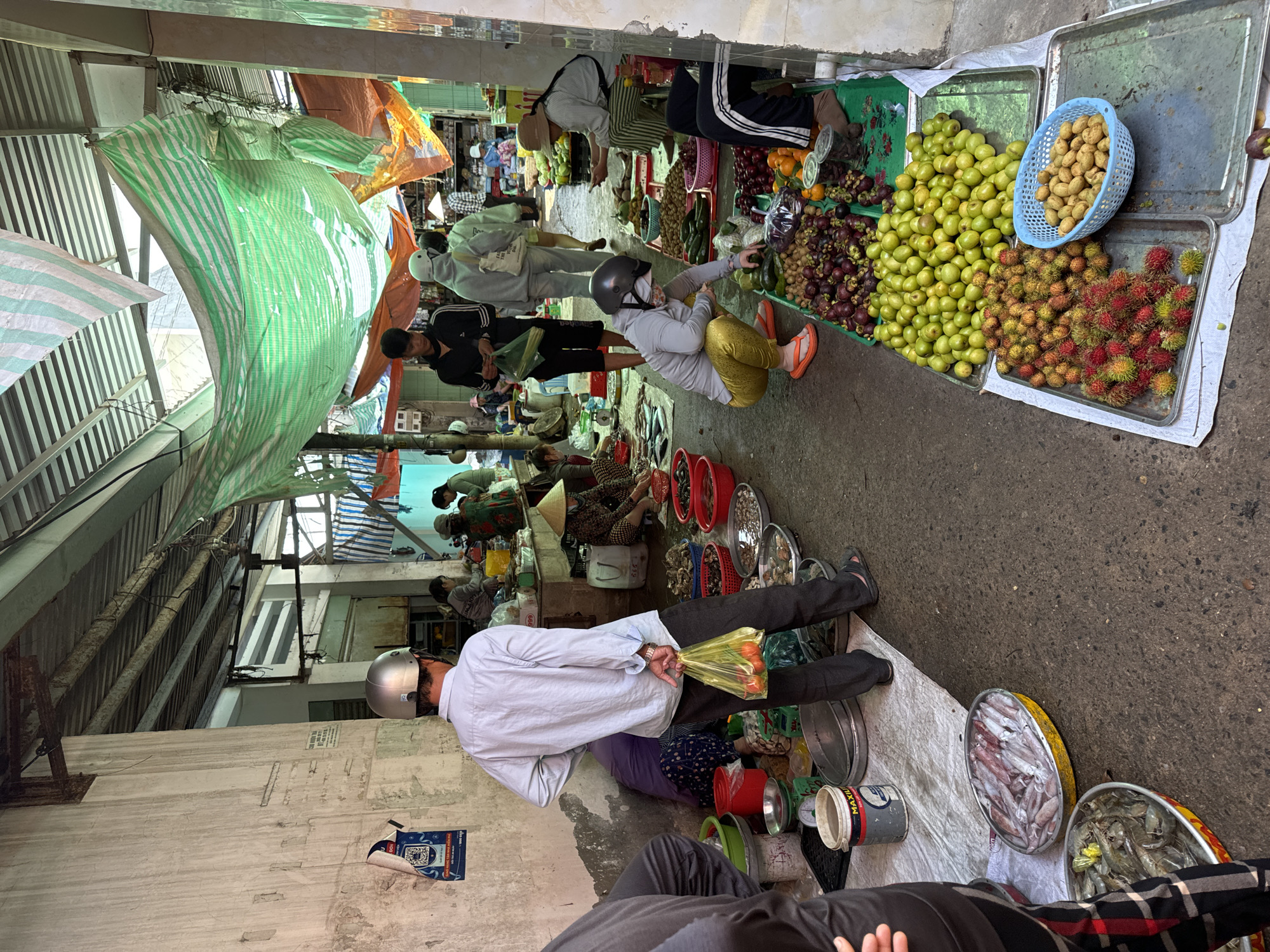 This image depicts an outdoor market scene in Mũi Né, Vietnam. The market is covered by a corrugated metal roof with various tarps and sheets of fabric strung up for shade. The ground is paved with asphalt and appears to be wet in some areas.
In the foreground, a person is partially visible on the left, wearing dark pants with red and black plaid stripes. To their right, a large oval metal pan is filled with squid. Below this pan, another similar pan contains shrimp. A large white tarp is spread out on the ground, on which various items are placed.
Moving further into the market, a man wearing a light blue long-sleeved shirt, dark pants, and a silver helmet stands with his back to the camera, holding a yellow plastic bag containing red fruits. To his left, a woman in a purple top and dark patterned pants is seated on the ground. Various containers of seafood, including shellfish and clams, are displayed in shallow metal pans and red plastic tubs on the ground.
Further back, several stalls are visible with vendors and customers interacting. One vendor, wearing a conical hat, is preparing food. Another person wearing a white helmet and gray jacket is examining produce. Numerous piles of fresh fruits and vegetables are on display, including green citrus fruits, rambutan, and mangosteen. Several people are seen shopping and moving through the crowded market. The overall impression is a busy, vibrant local market.