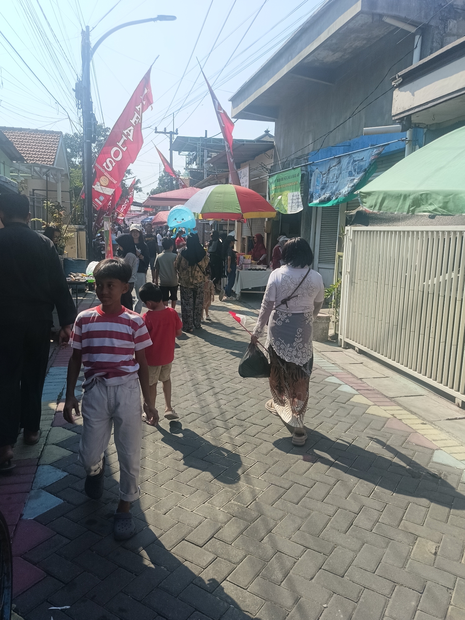 The image depicts a street scene in Gedangan, Indonesia, with people walking on a paved sidewalk. The sky is clear and bright, suggesting a sunny day. Red flags with white lettering are visible, hanging from poles on the left side of the street. Several people, including men, women, and children, are present, some walking individually and others in groups. They are dressed in casual clothing. To the right of the sidewalk, there are buildings and stalls, some with awnings. A white fence runs along the right side of the sidewalk. The ground is paved with interlocking bricks, and shadows of people and objects are cast on the ground, indicating the direction of the sunlight. In the foreground, two boys are walking, with one wearing a striped shirt and light-colored pants, and the other in a red shirt and shorts. A woman in a patterned skirt and white top is walking away from the camera on the right side of the sidewalk, carrying a black bag. The scene appears to be a lively street with pedestrians and vendors.