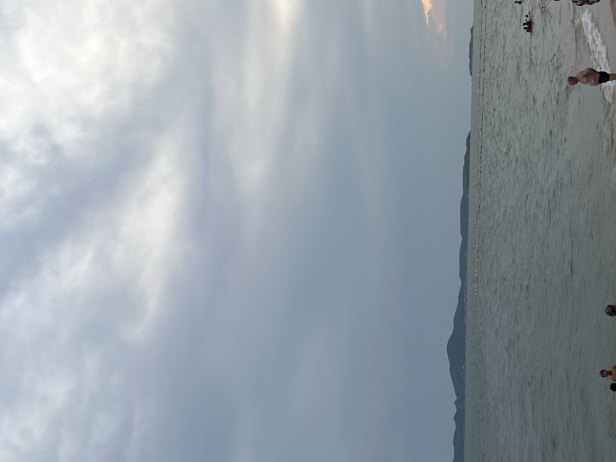 The image depicts a beach scene, likely in Pattaya City, Thailand. The view is predominantly of the sky, which is overcast with gray and white clouds. To the right, there's a strip of land with a mountainous or hilly silhouette. The ocean occupies a portion of the right side, with individuals wading or swimming in the water. Some are closer to the shore, while others are further out. The water is a murky gray color, and there are a few indistinct figures on the beach near the water's edge.