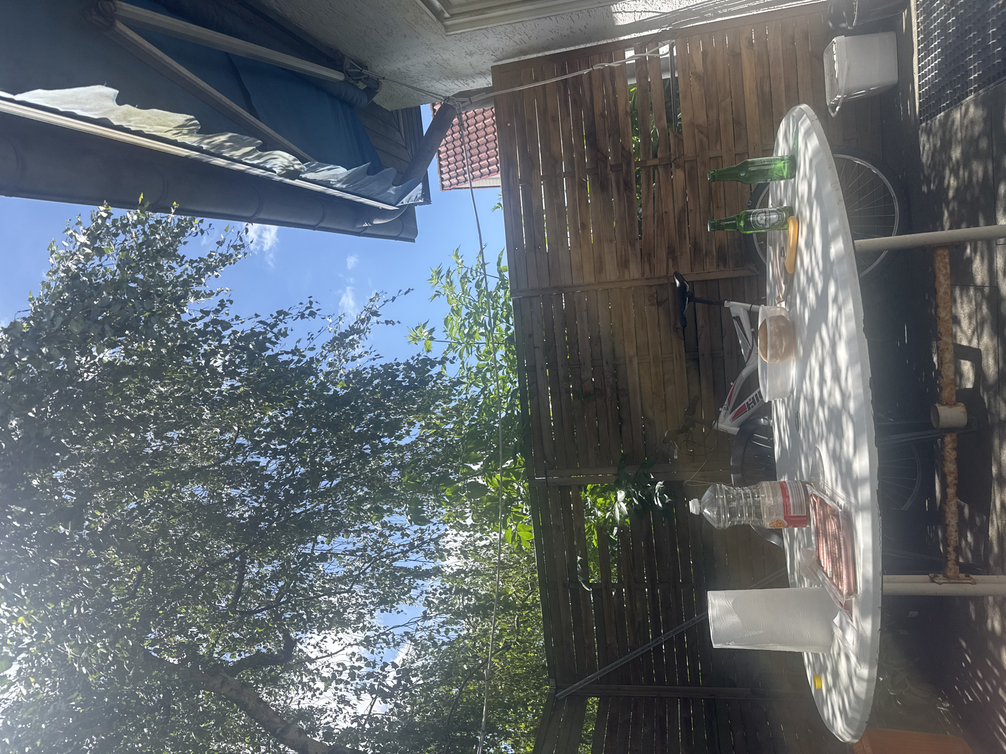 The scene depicts an outdoor patio area in Hattersheim am Main, Germany, under bright sunlight on a clear day with scattered cumulus clouds. No individuals are visible.
In the foreground, a white round outdoor table with a white metal base is positioned centrally. On the table, from left to right, are a vertical roll of white paper towels, a clear plastic bottle with an orange-and-white label (partially visible, resembling Aldi branding), a vacuum-sealed package containing several frankfurters or similar sausages, a white plastic container holding several light-colored bread buns, a yellow object (possibly a kitchen tool or handle), and two green glass bottles, one clearly labeled "Heineken." Shadows from tree leaves and possibly nearby structures are cast across the table and the ground beneath.
Behind the table, a white bicycle with a dark seat and handlebars is leaning against a horizontal slatted wooden fence. The fence comprises multiple horizontal wooden planks of varying shades of brown. A light-colored cable or hose runs horizontally across the top section of the fence.
To the right of the table and bicycle, on the ground, a white cooler box is situated next to a dark bucket. Further right, a metal grate or drain cover is visible on the paved ground.
The background consists of dense foliage from a large deciduous tree, likely a birch, particularly prominent in the upper left, casting mottled light and shadow. Beyond the fence to the right, a multi-story building with a light-colored stucco-like exterior is visible. Attached to this building is a dark grey rain gutter and a partially furled blue fabric awning. Above the fence and to the right, a glimpse of a red-tiled roof from another distant structure is visible against the blue sky.