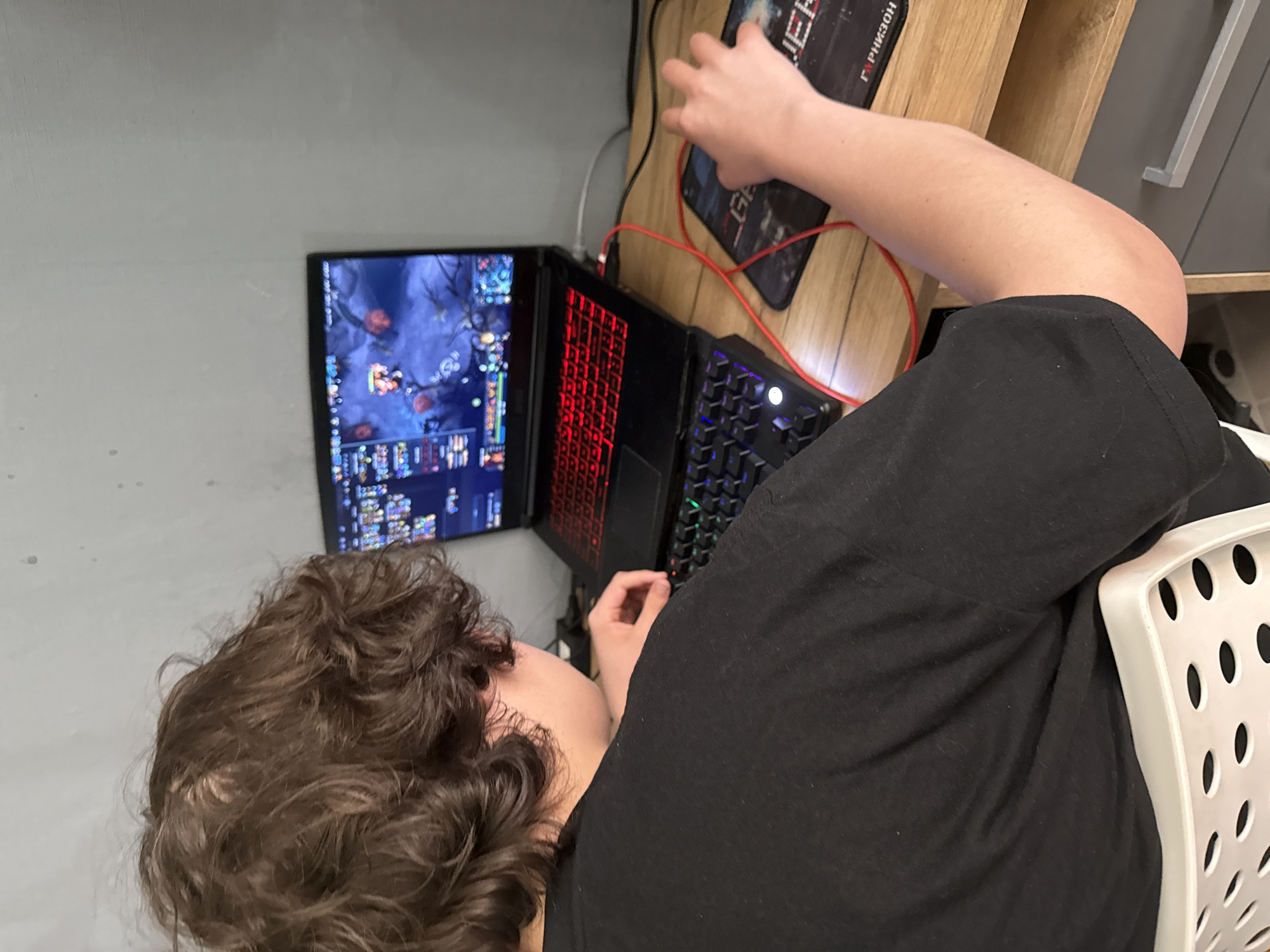 A person with curly brown hair is seated in a white chair, using a gaming laptop and an external keyboard. The laptop screen displays a game with a blue and purple interface. A black mousepad with white lettering and a logo is positioned on a wooden surface next to the laptop and keyboard. The person is wearing a black shirt. The environment includes a gray wall and a light brown cabinet with a white door. The location is not discernible from the provided image.