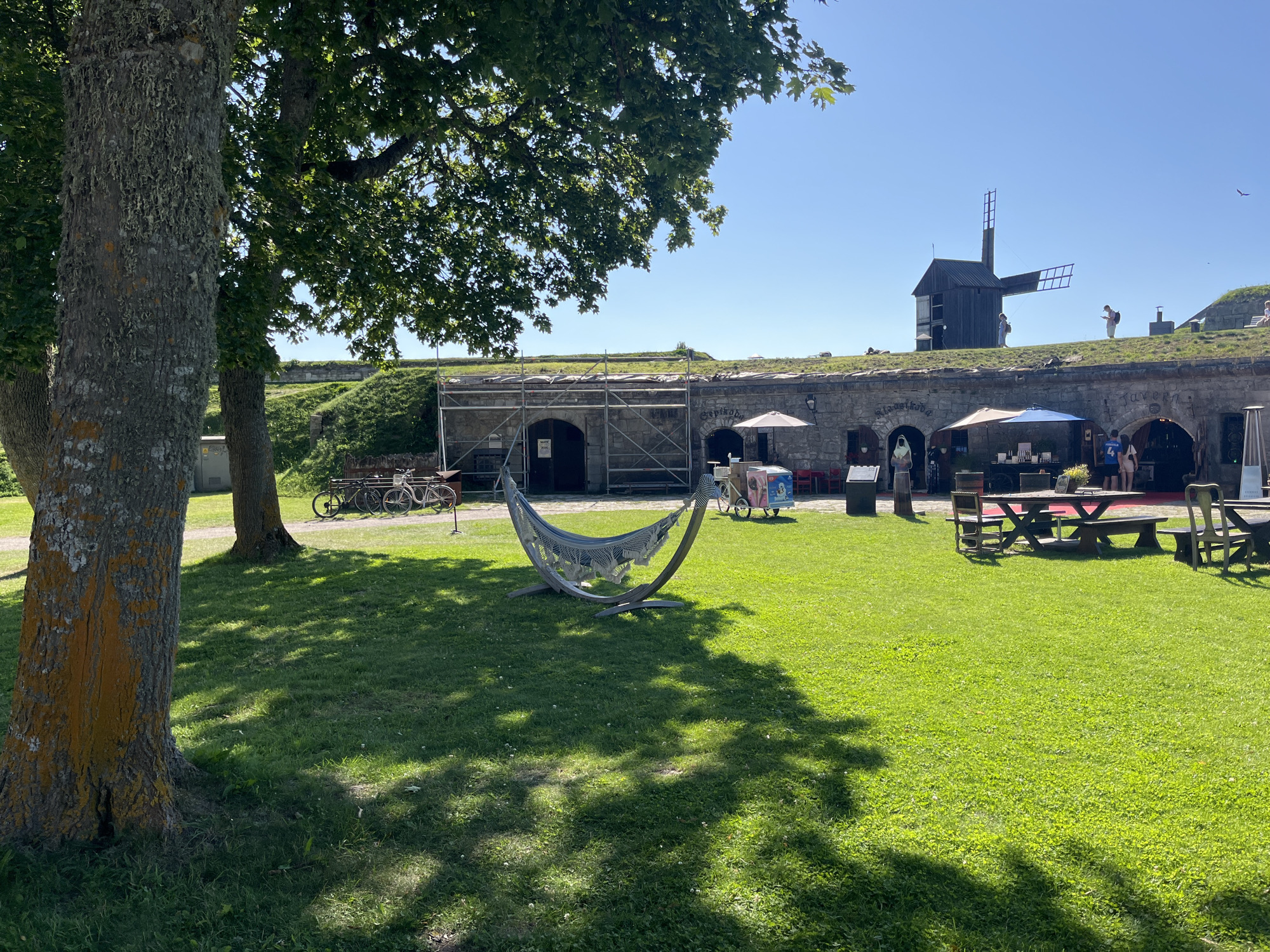 The image depicts a sunny outdoor scene at Kuressaare Castle in Kuressaare, Estonia. In the foreground, a hammock made of white netting is suspended on a wooden frame, situated on a lush green lawn. Several trees with dappled sunlight filtering through their leaves cast shadows across the grass. To the left, the textured bark of a large tree with orange lichen is prominent.
In the background, a stone fortress wall forms a semicircular structure. Several archways are visible in the wall, some with scaffolding in front of them, suggesting ongoing construction or maintenance. Outdoor seating areas with tables and umbrellas are set up in front of some of these archways, indicating a place for dining or relaxation.
Above the fortress wall, a traditional wooden windmill with two sails stands on a grassy mound. People are visible walking on the mound near the windmill. The sky is clear and blue.
In the middle ground, several bicycles are parked near one of the archways. An ice cream cart is also visible. A few people are standing and interacting in front of the archways, some appearing to be customers or staff. The overall atmosphere is one of a pleasant, historical tourist attraction on a bright day.