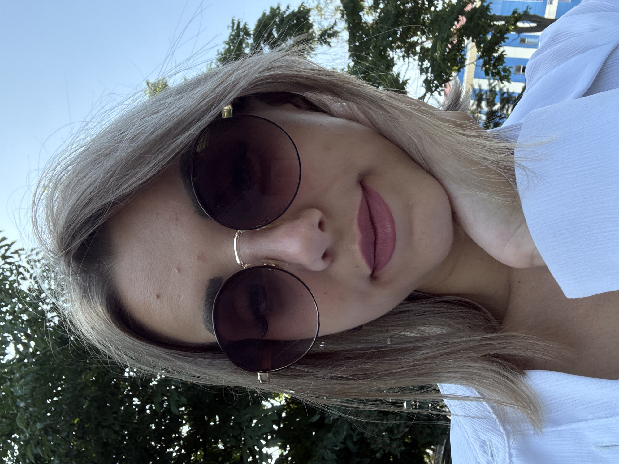 A young woman with blonde hair is taking a selfie outdoors. She is wearing large, round sunglasses with dark lenses and a gold frame, and a white collared shirt. Her left hand is resting on her chin. Behind her, a tree with green leaves is visible against a bright blue sky. Further in the background, a blue and white building can be seen. The image is likely taken in Yuzhno-Sakhalinsk, Russia, given the building's architecture.