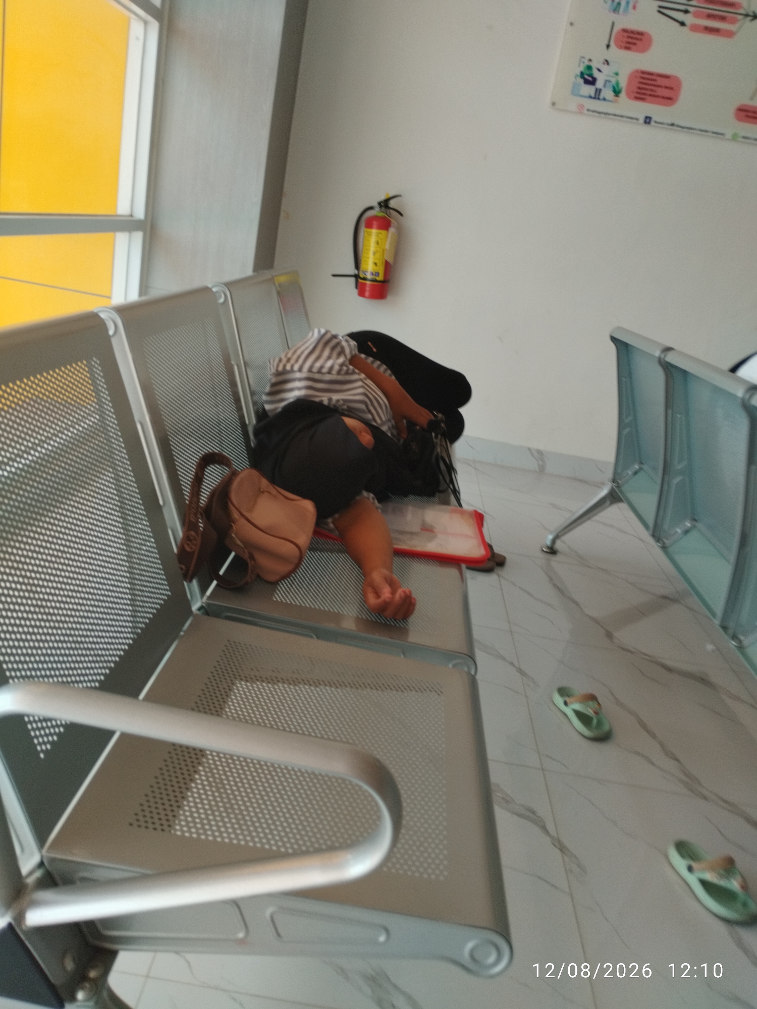 A person is sleeping on a bench in a waiting area, likely a hospital or office building. They are lying on their side with their head resting on their arm. A brown purse is placed on the bench next to them. There is a fire extinguisher mounted on the wall behind them, and a colorful diagram is also on the wall. The floor is tiled with a marble pattern. Two pairs of green sandals are visible on the floor. The scene is lit by natural light coming through a large window with a yellow wall visible behind it. The date stamp on the image reads "12/08/2026 12:10". The location is identified as Bandar Lampung, Indonesia.