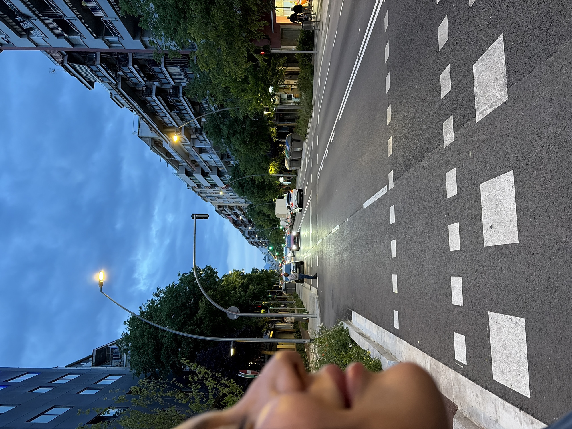 The image depicts a street in Granollers, Spain, likely taken during the early evening. The perspective is ground-level, slightly angled, with part of a person's arm and shoulder visible in the foreground. The street is multi-lane, paved with dark asphalt, and marked with white lines and square blocks. The lanes appear to be directional. Buildings line both sides of the street. The buildings on the left are taller, multistory structures with visible balconies. Trees are planted along the sidewalk. Streetlights are present, illuminating the area. Several cars are visible further down the street, with headlights on, suggesting twilight conditions. A person stands on the sidewalk near a bin. The sky is blue with cloud cover.