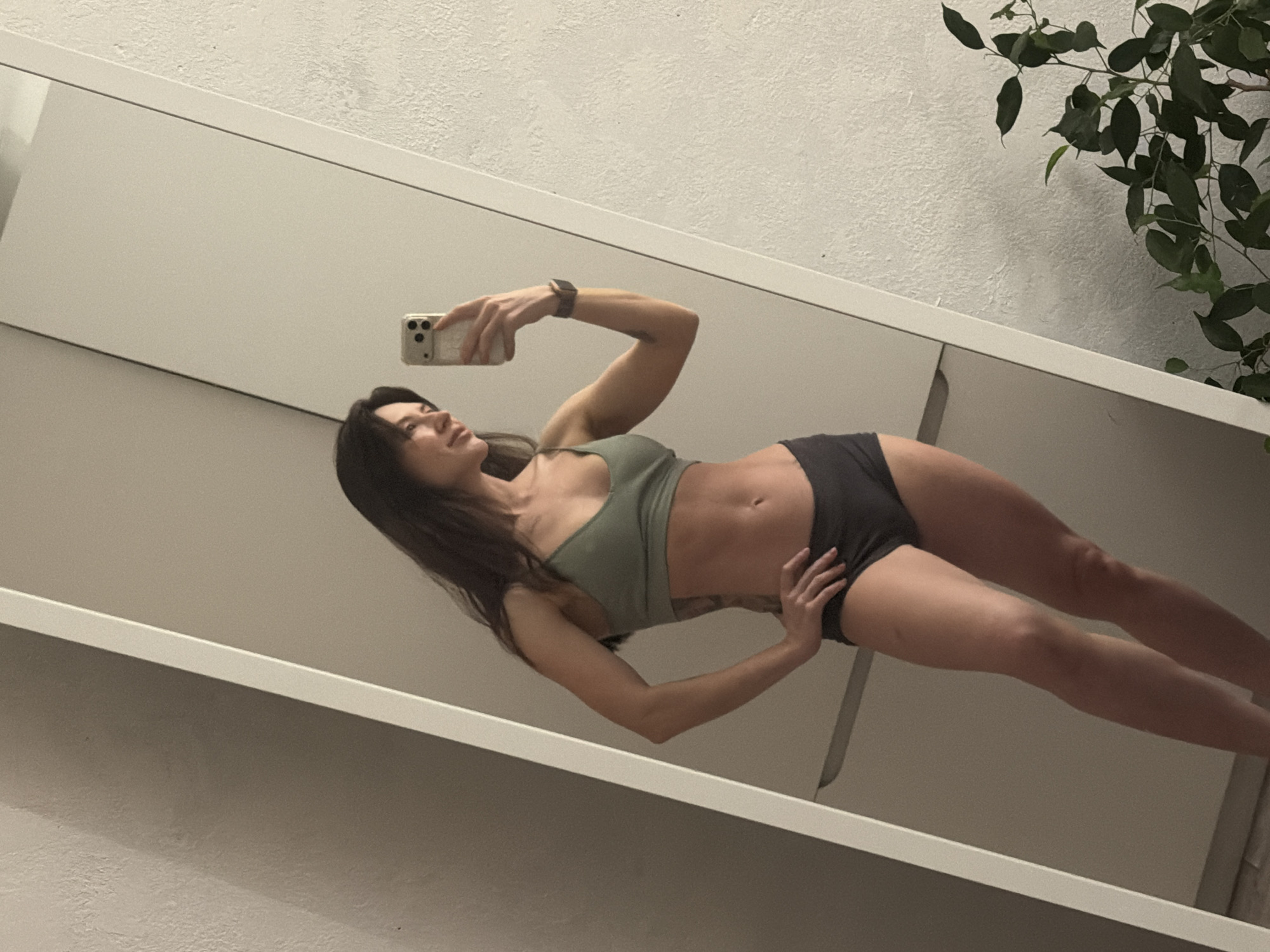 A woman is posing in a mirror selfie in what appears to be a bathroom. She is wearing a green sports bra and gray underwear, and holding a smartphone in her right hand. The background shows a white wall and a plant. The location is not identifiable as Perm, Russia, as there are no distinguishable landmarks.