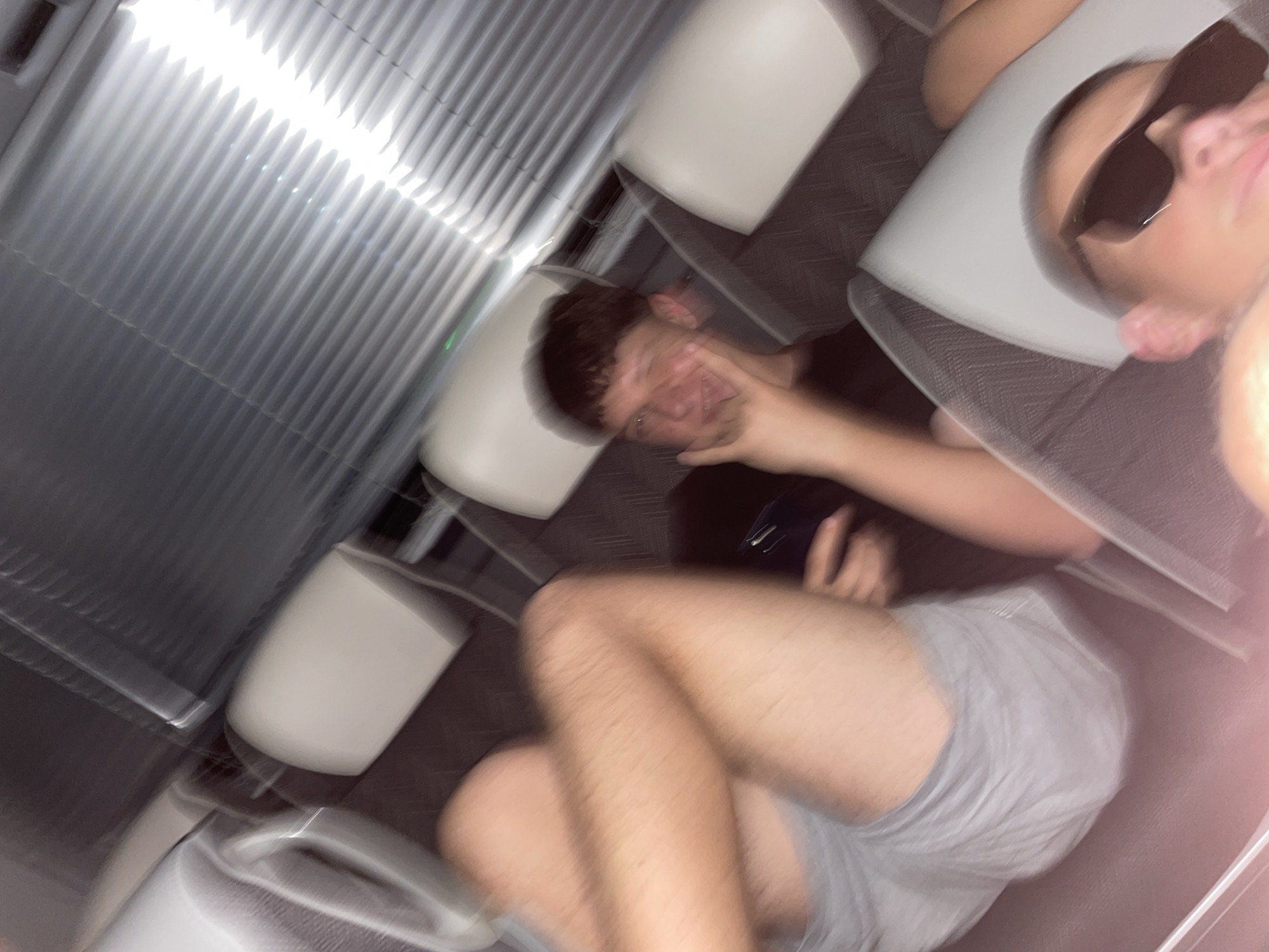 The image shows the interior of a train or airplane, with rows of white seats. Two people are visible. One person is wearing sunglasses and is partially visible in the upper right corner. Another person is seated with their hand near their face. This person is wearing a black shirt. The seated person is also wearing gray shorts. The environment appears to be clean, but the image is blurry due to motion or camera shake. The location in the city of Shtarkovo, Bulgaria, cannot be confirmed from the image.