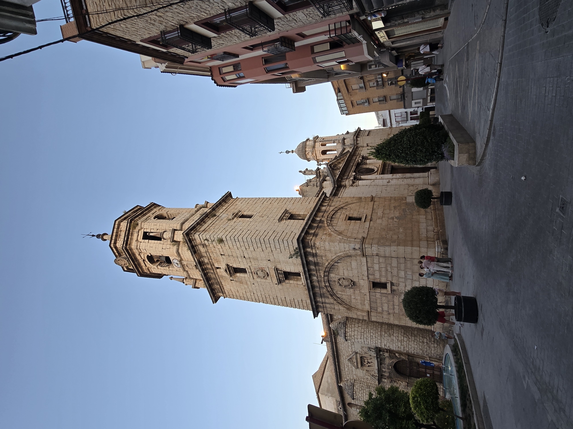 The image depicts a street scene in Jaén, Spain, dominated by the imposing facade of the Jaén Cathedral and its bell tower. The architecture is distinctly baroque, with the sandstone structure of the cathedral featuring arched windows, decorative stonework, and a prominent clock face on the tower. To the right of the cathedral, a narrow street curves away, lined with multi-story buildings with balconies and shops at street level.
Several people are present in the scene. In the foreground, near a fountain and a small ornamental tree, a group of individuals are standing and conversing. Further down the street, other pedestrians are walking, some appearing to be tourists. The street itself is paved with cobblestones, and the setting suggests it is late afternoon or early evening, with the sky a clear, pale blue.
The overall impression is of a historic urban environment, with the cathedral serving as a central landmark. The lighting is soft, casting gentle shadows, and the atmosphere appears calm and peaceful. The image captures a slice of daily life in a Spanish city, blending religious architecture with commercial activity and pedestrian movement.