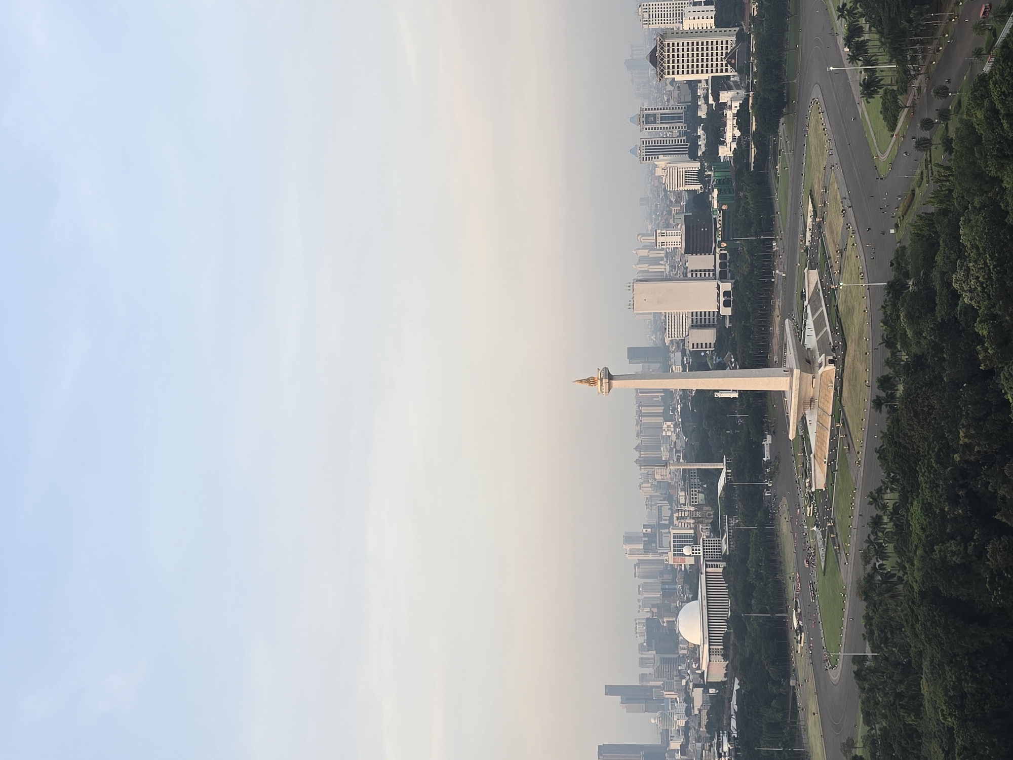 The image shows an aerial view of Central Jakarta, Indonesia. A tall monument, likely the National Monument (Monas), stands prominently in the scene. Buildings and skyscrapers surround the monument, and roads with vehicular traffic can be seen. Trees and green spaces are also visible. The sky is overcast, with a hazy quality to the air.