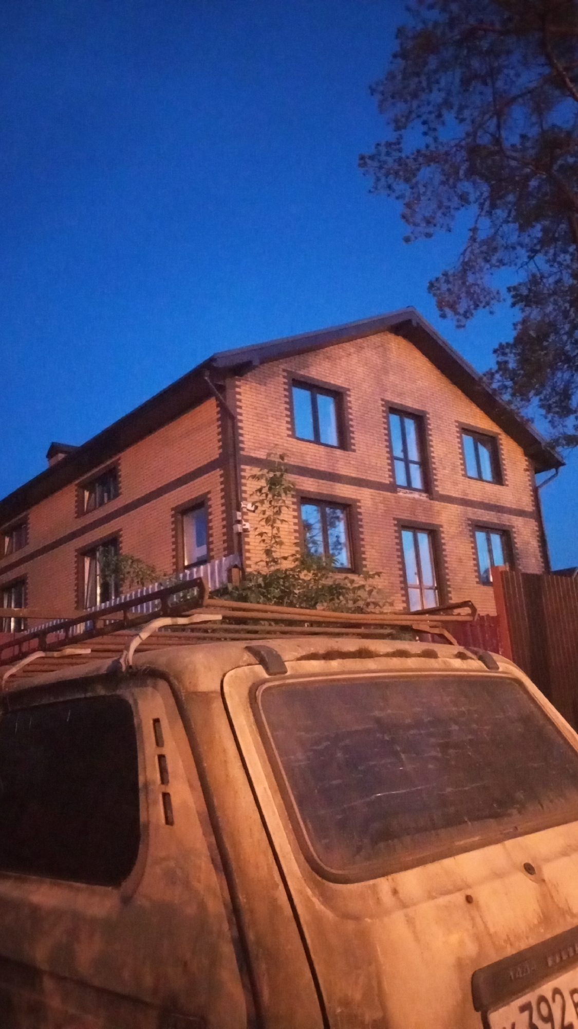 A nighttime view in Юдино, Russia, shows a two-story brick house with a dark brown roof and window frames. The house has a symmetrical facade with three windows on each floor. In the foreground, an old, light-colored car with a roof rack obscures the lower portion of the house. A small tree or shrub grows near the car and the house's facade. A wooden fence is visible to the right of the house, and a tree with sparse leaves stands in the upper right corner. The sky is a deep blue, suggesting it is either dusk or dawn. The car's license plate is partially visible and appears to have Russian characters and numbers.