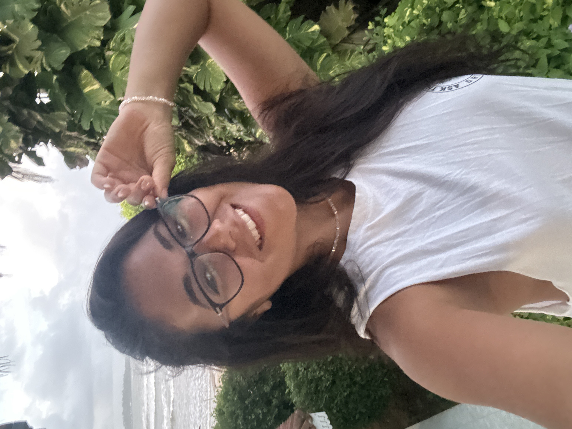 A young woman with long, dark hair and glasses smiles at the camera in a selfie. She is wearing a white sleeveless shirt and a delicate bracelet on her wrist. The background shows lush green foliage and the ocean with waves. The image appears to be taken outdoors in a tropical location. The specific location in Peleana, Sri Lanka, is not identifiable from the visual information.
