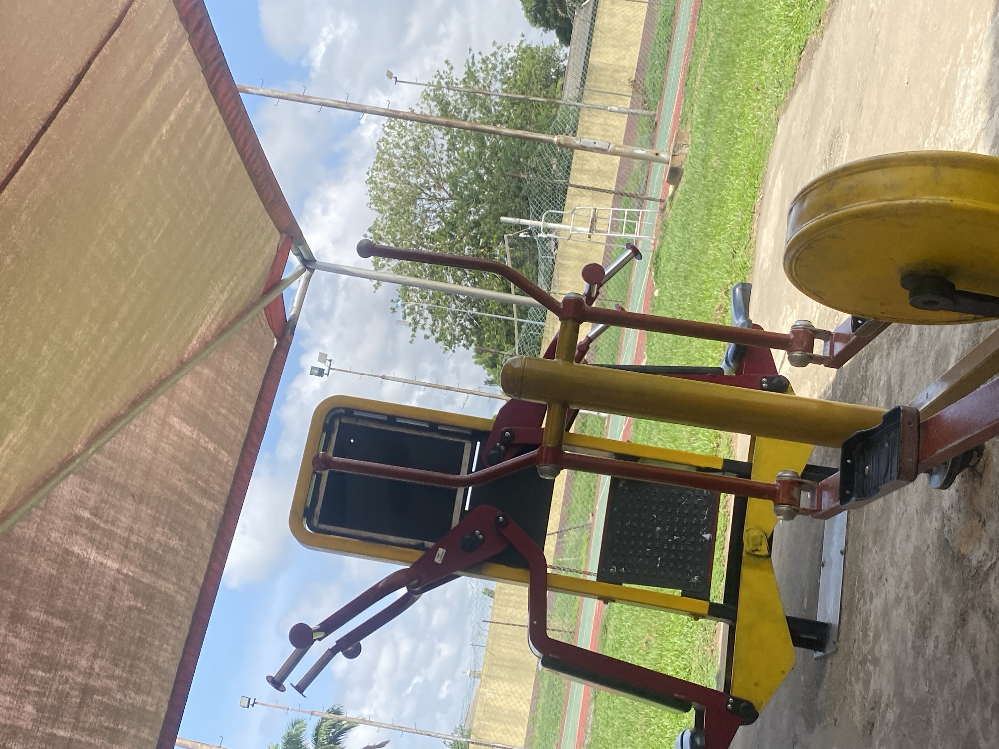 A stationary exercise machine occupies a park or public fitness area in Ikorodu, Nigeria. The equipment is constructed with red and yellow-painted metal. The scene is in an outdoor setting with bright sunlight and blue sky with puffy clouds overhead. The surroundings consist of grass, trees, and a mesh-wire fence. A beige canopy or sunshade is visible in the upper left frame corner.