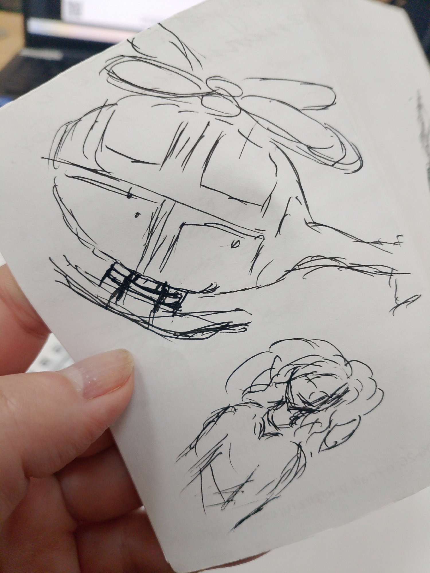 The image shows a hand holding a piece of paper with pen drawings of a helicopter and a person with curly hair. The background is blurred. There is no information in the image to determine its location as Izhevsk, Russia.