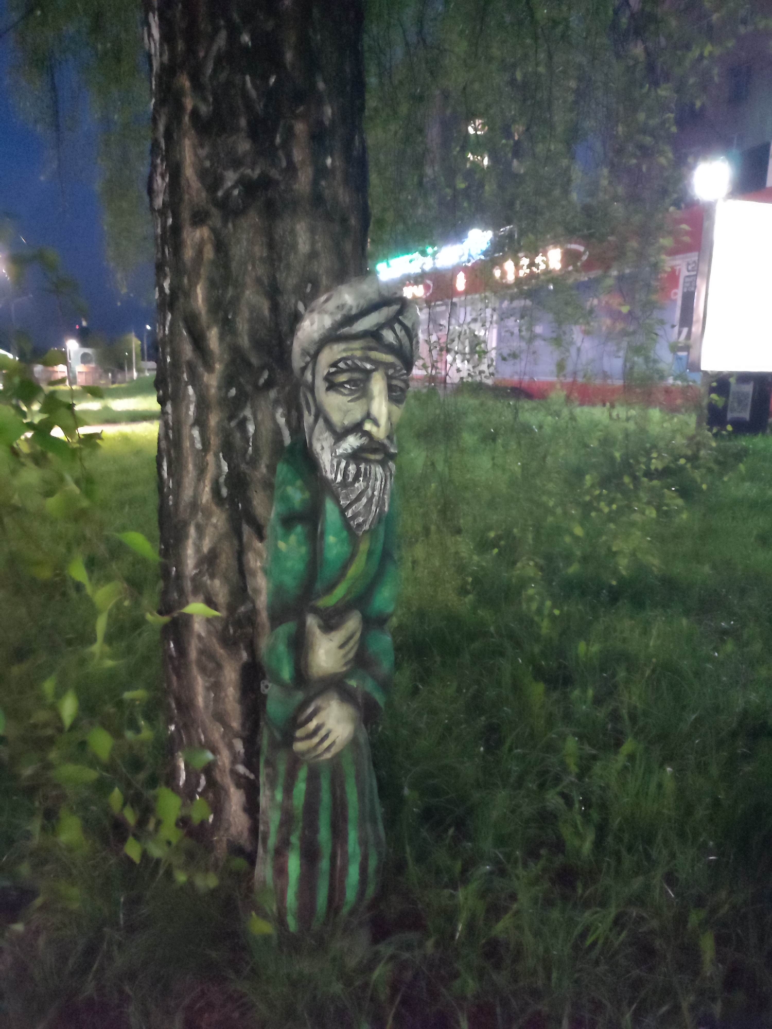 A carved, painted wooden figure of a man with a turban and beard stands in front of a tree trunk in a grassy area at night. The figure is dressed in a green robe with dark vertical stripes. Beyond the figure, the scene shows a grassy area with low vegetation and glimpses of buildings with lighted signs in the background. A bright billboard is also visible on the right side of the image. The location is Unknown, Unknown.