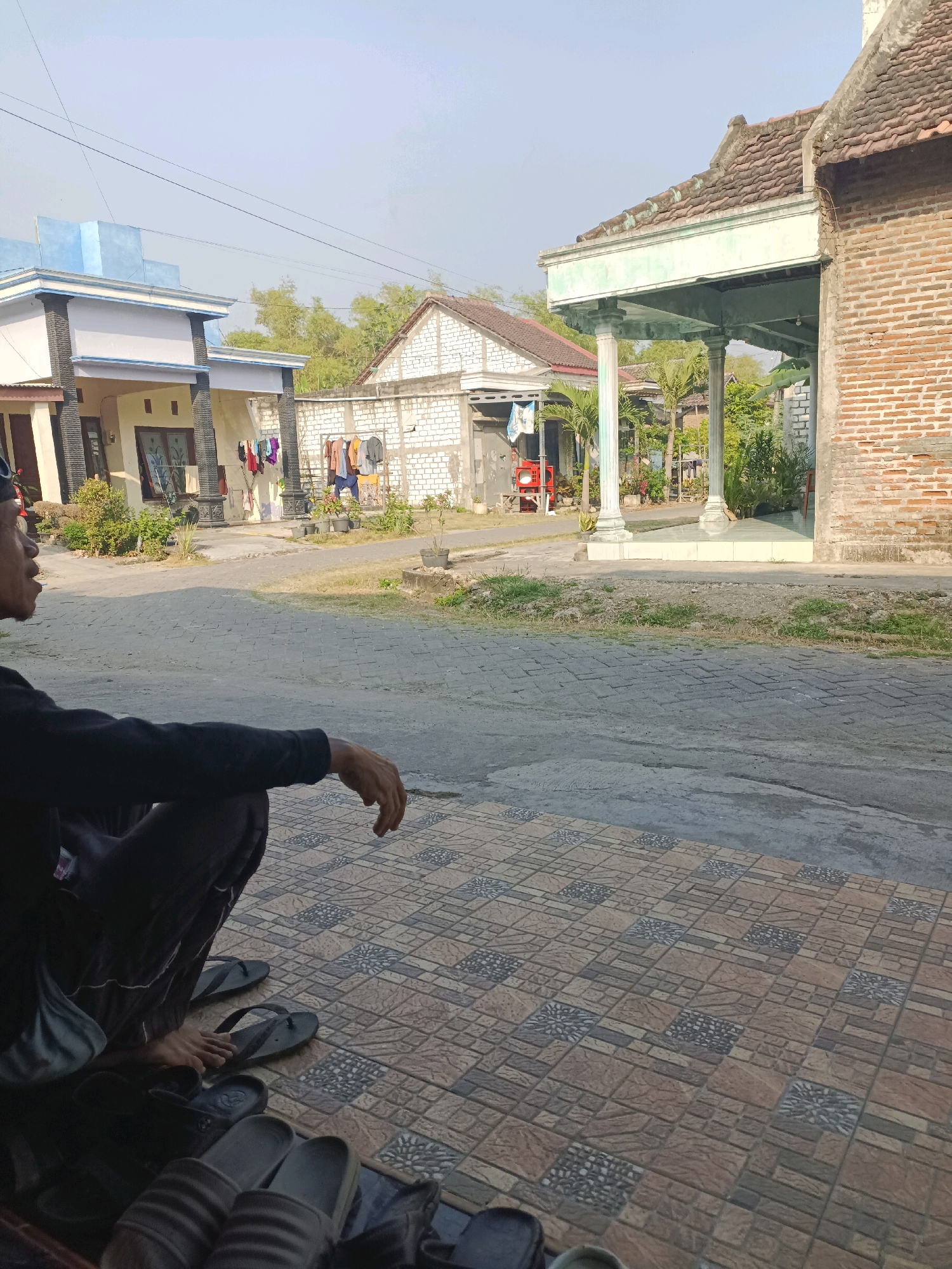 In Rancang Kencono, Indonesia, a person in a black long-sleeved shirt and dark pants sits on a tiled patio, looking to the left. Several pairs of black and grey flip-flops are on the patio near the person. Across a paved walkway, a red utility cart is visible next to a white brick wall. Further back, a house with light blue trim and a white facade stands under a clear sky. To the right, a brick building with a terracotta tiled roof is situated. Trees and other vegetation are scattered throughout the scene.
