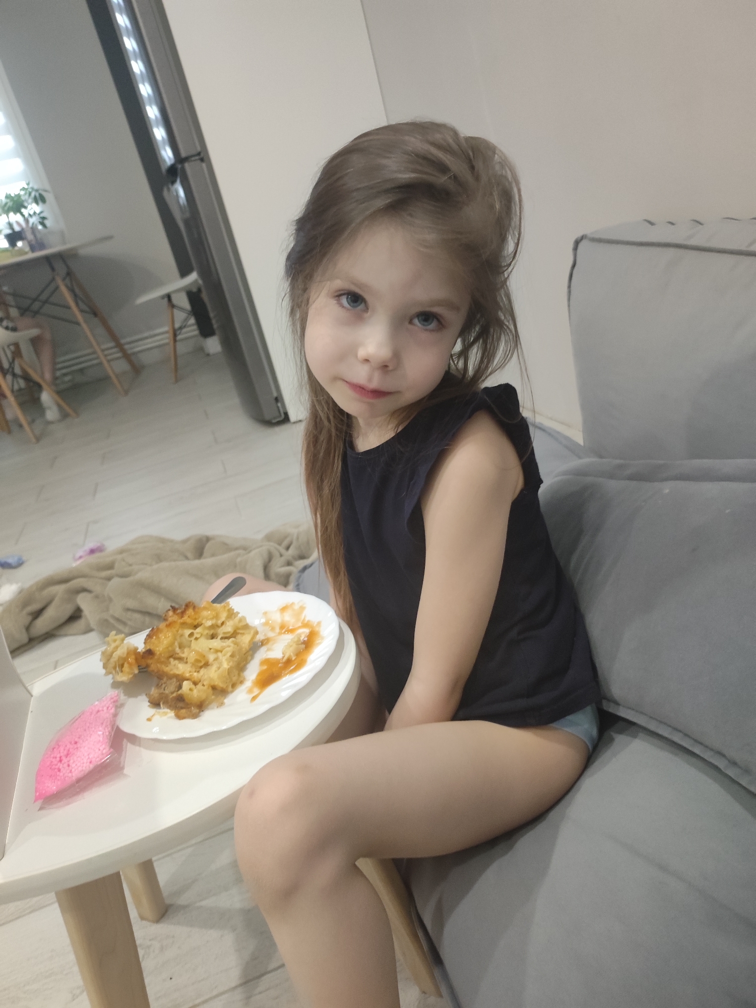 A young child with brown hair and blue eyes sits on a gray sofa next to a white table. A plate of food, mostly pasta, and a pink object are on the table. In the background, there's a kitchen area with white furniture and a window. The location appears to be inside a home, possibly in Votkinsk, Russia, but this cannot be confirmed from the image alone.