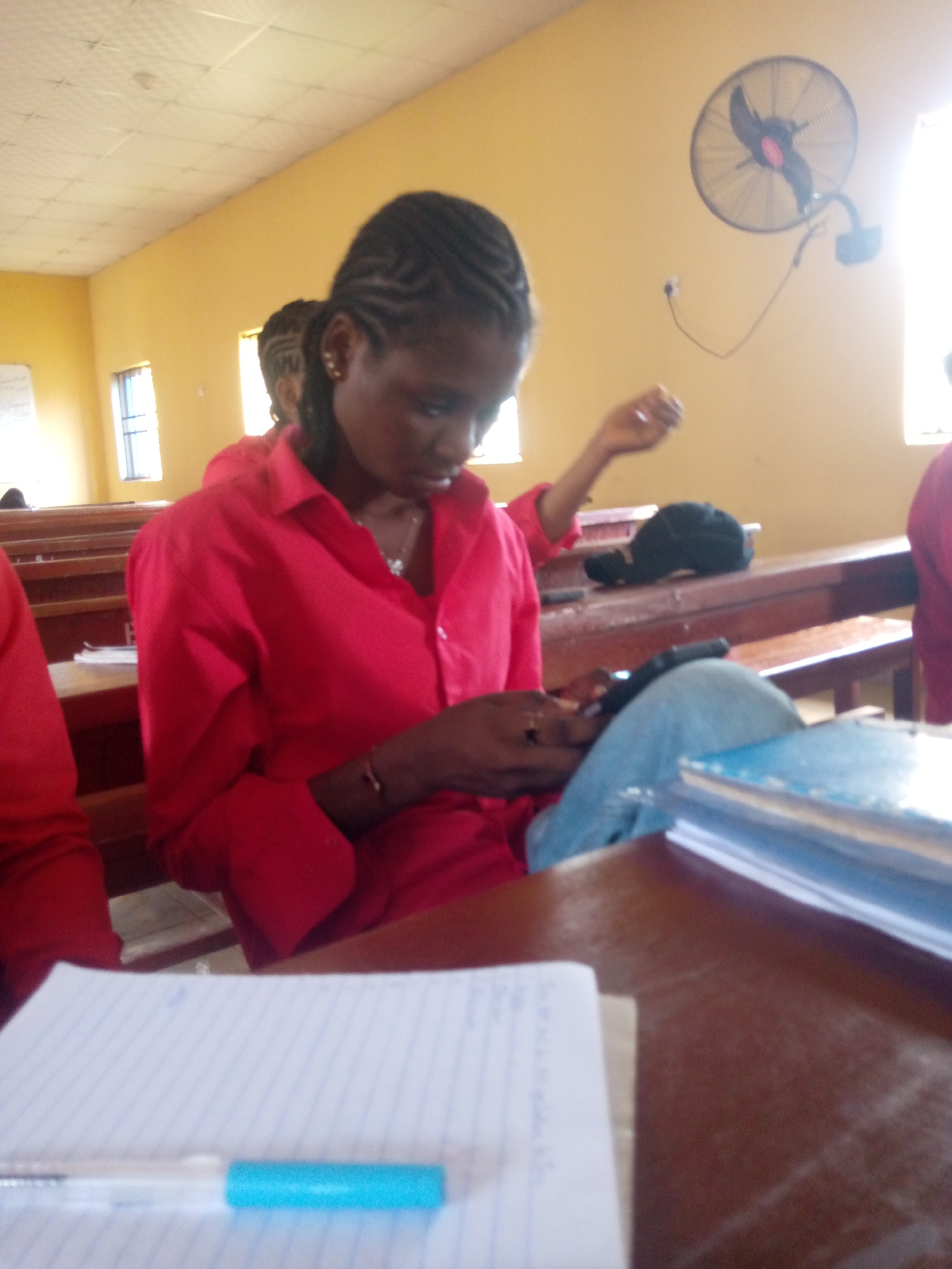The image depicts a classroom scene in Nsugbe, Nigeria, with a young Black woman in the foreground. She is wearing a bright red collared shirt and appears to be focused on her mobile phone, which she holds in her hands. Her hair is styled in cornrows. The setting is a classroom with wooden desks and benches. In the background, another student can be seen wearing a similar red shirt, suggesting a uniform or a school event. Sunlight streams in from a window, illuminating the room and creating bright spots on the walls. A wall-mounted fan is visible on the yellow-painted wall, indicating the need for cooling in what is likely a warm climate. Papers and books are scattered on the desks, suggesting a typical school day with learning activities. The lighting suggests it is daytime. There is no visible text in the image. The overall mood appears to be one of quiet concentration and everyday classroom activity.