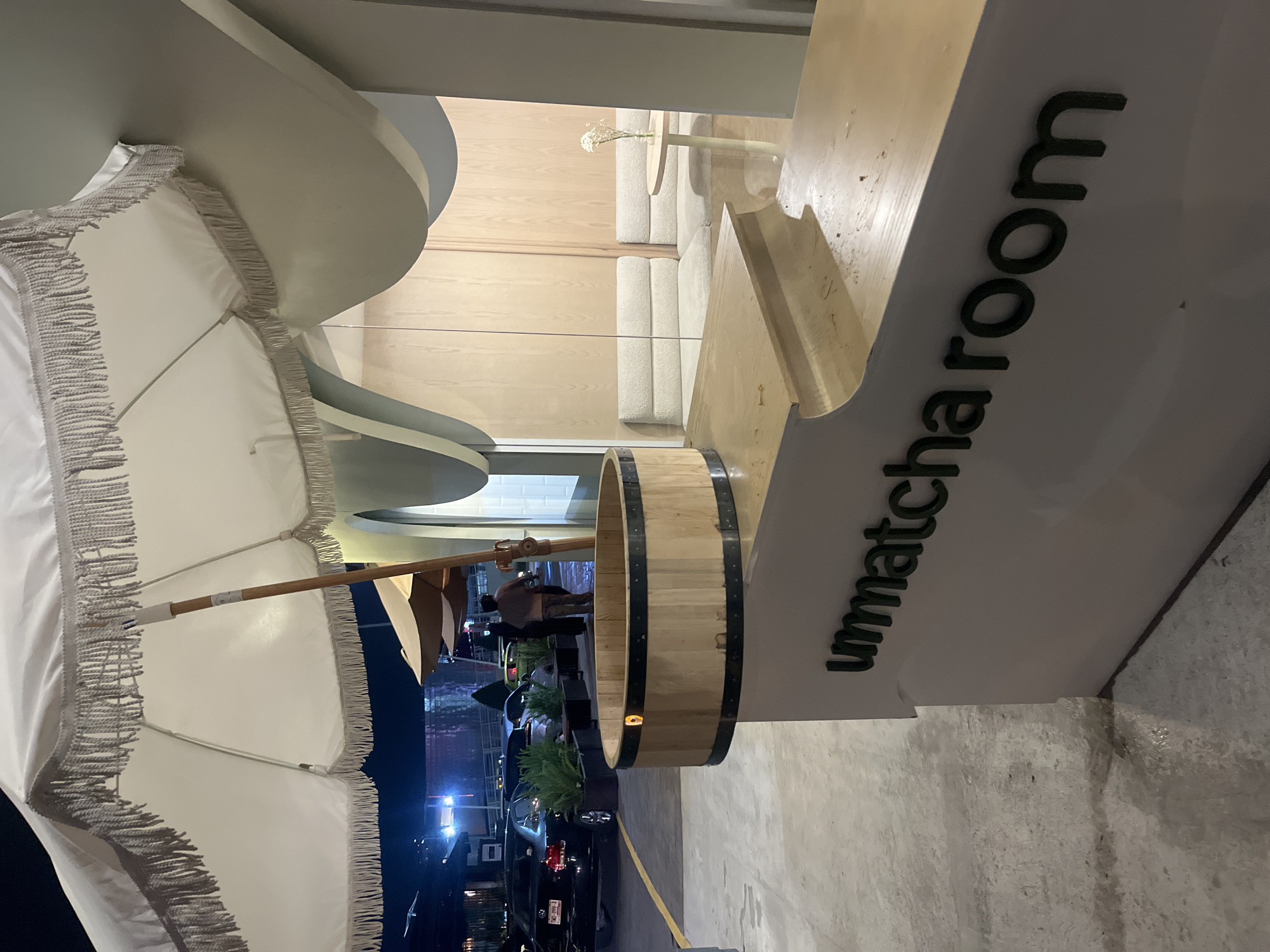 This nighttime, street-level view captures "unmatcha room," as printed on a sign, potentially the name of a restaurant in Puebla, Mexico. The environment consists of an outdoor dining space with white umbrellas, whose fringe and wooden structure are visible. Inside, the restaurant features minimalist, light-colored wood and seating. A wooden barrel planter appears near the restaurant's entrance, and a city street is visible beyond the dining area, with cars parked along the curb. Dimly lit streetlights are seen. At least two individuals are visible in the dining area, though their actions or interactions cannot be discerned from this perspective.