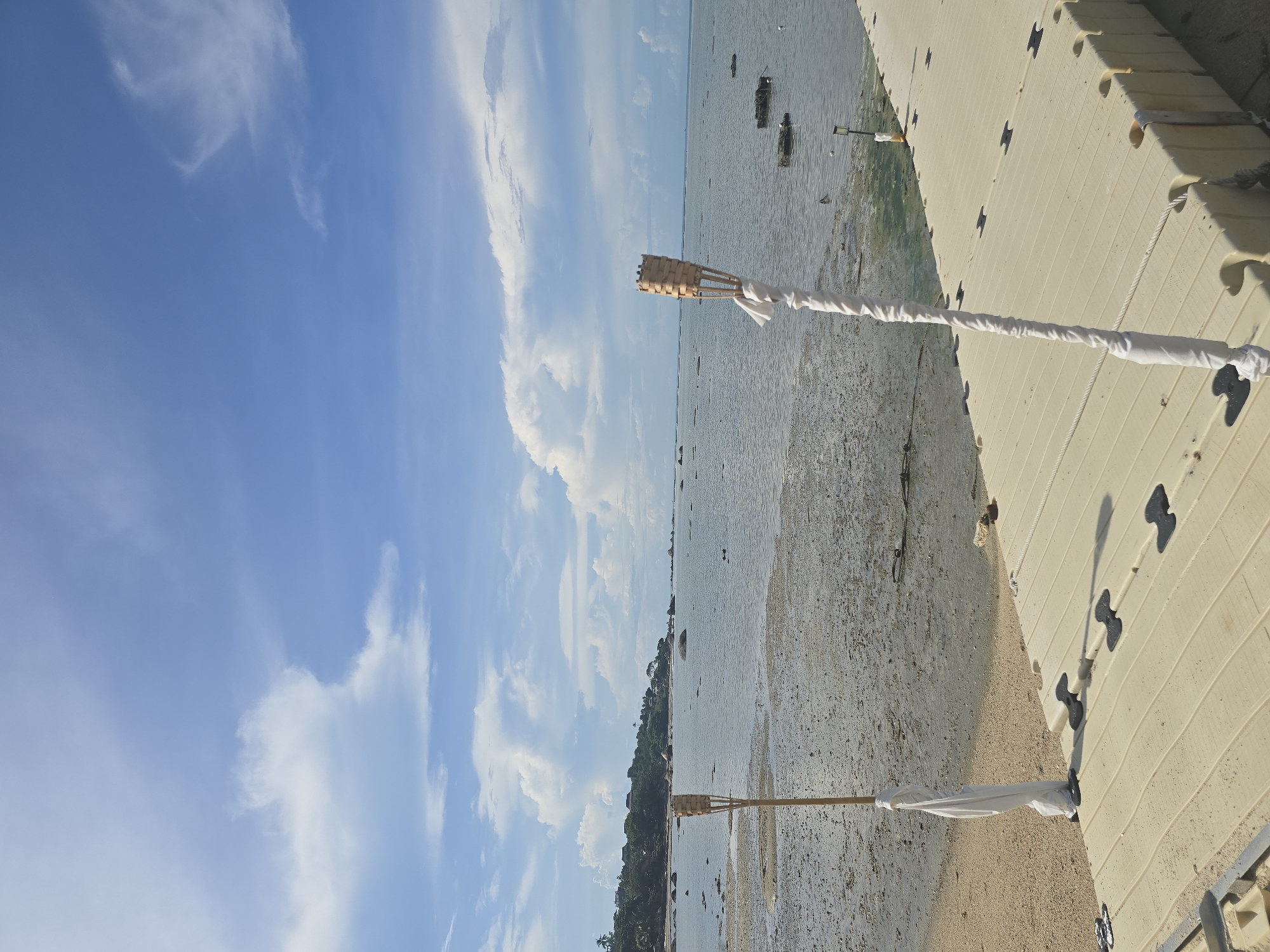 A bright, daytime, eye-level shot of a beach at low tide in Baan Chaweng Noi, Thailand. In the foreground, a light-colored plastic walkway extends from the right side of the frame towards the left, with two tiki torches positioned on the walkway. The torch on the left is partially obscured by the walkway, while the torch on the right stands prominently. The walkway is a light tan color and appears to be made of interlocking modular sections. Both torches are bamboo with woven tops, and their poles are wrapped in white fabric. The ground transitions from the sandy beach to the left of the walkway to the muddy, shallow water and exposed seabed to the right. The water is calm and reflects the blue sky with scattered white clouds. In the distance, a tree-covered hillside with several buildings is visible along the coastline, with rocks jutting out of the water. The sky is a vibrant blue with a mix of large, puffy white clouds and wispy cirrus clouds. There are no people visible in the scene.