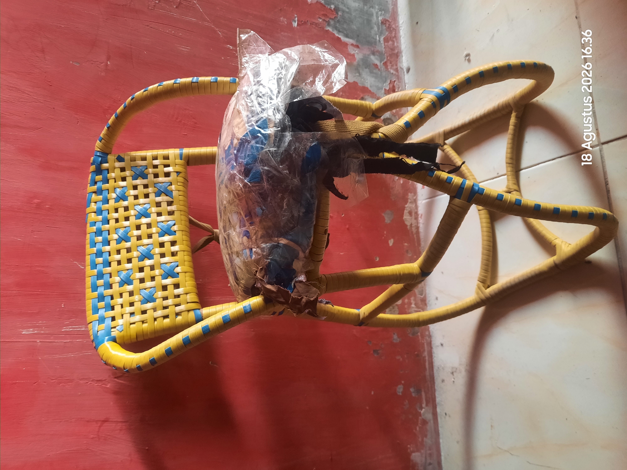 A yellow woven chair with blue accents stands against a red wall. The chair has a woven backrest and frame. The seat of the chair is covered with a clear plastic sheet that is partially torn, revealing brown fabric and what appears to be padding underneath. The chair is on a tiled floor, and a shadow is cast on the floor to its left. The background wall is textured and shows signs of wear, with peeling paint in some areas. A timestamp, "18 Agustus 2026 16.36," is visible in the bottom right corner, suggesting this image was taken in Sukokerto, Indonesia.