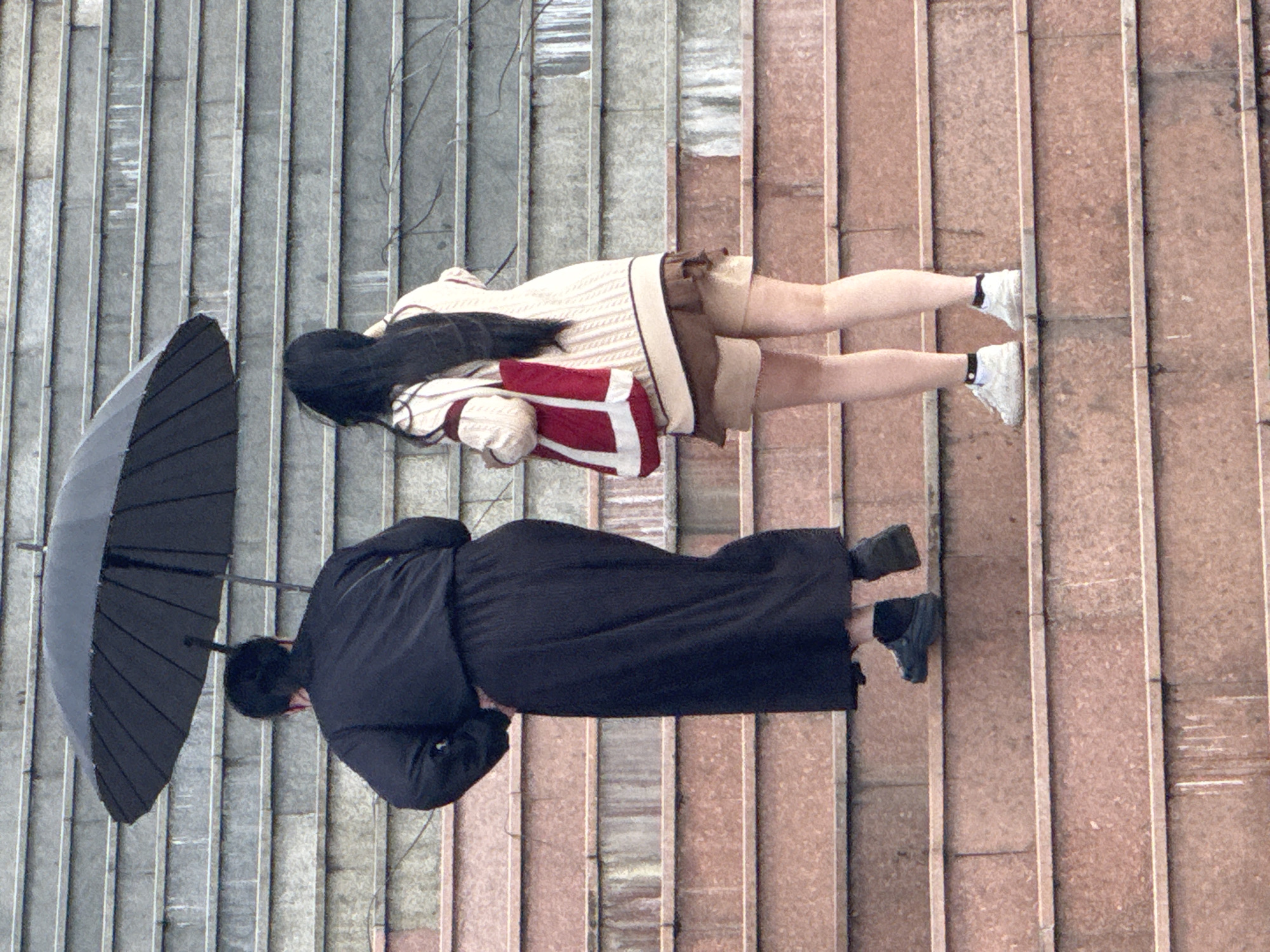Two individuals, viewed from behind, ascend a broad, outdoor staircase with stone steps. The person on the left, wearing a long black skirt and dark shoes, holds a large black umbrella over both of them. The person on the right, dressed in a cream-colored knit sweater, a brown skirt, and white sneakers, carries a red and white tote bag. The staircase appears to be a public feature, possibly part of a park or city infrastructure, with a gray, textured surface forming the risers of the steps. The lighting suggests an overcast or rainy day. The context of Da Lat, Vietnam, implies this scene is likely within the city's characteristic hilly terrain, known for its many staircases and lush surroundings, though these elements are not explicitly visible in the frame.