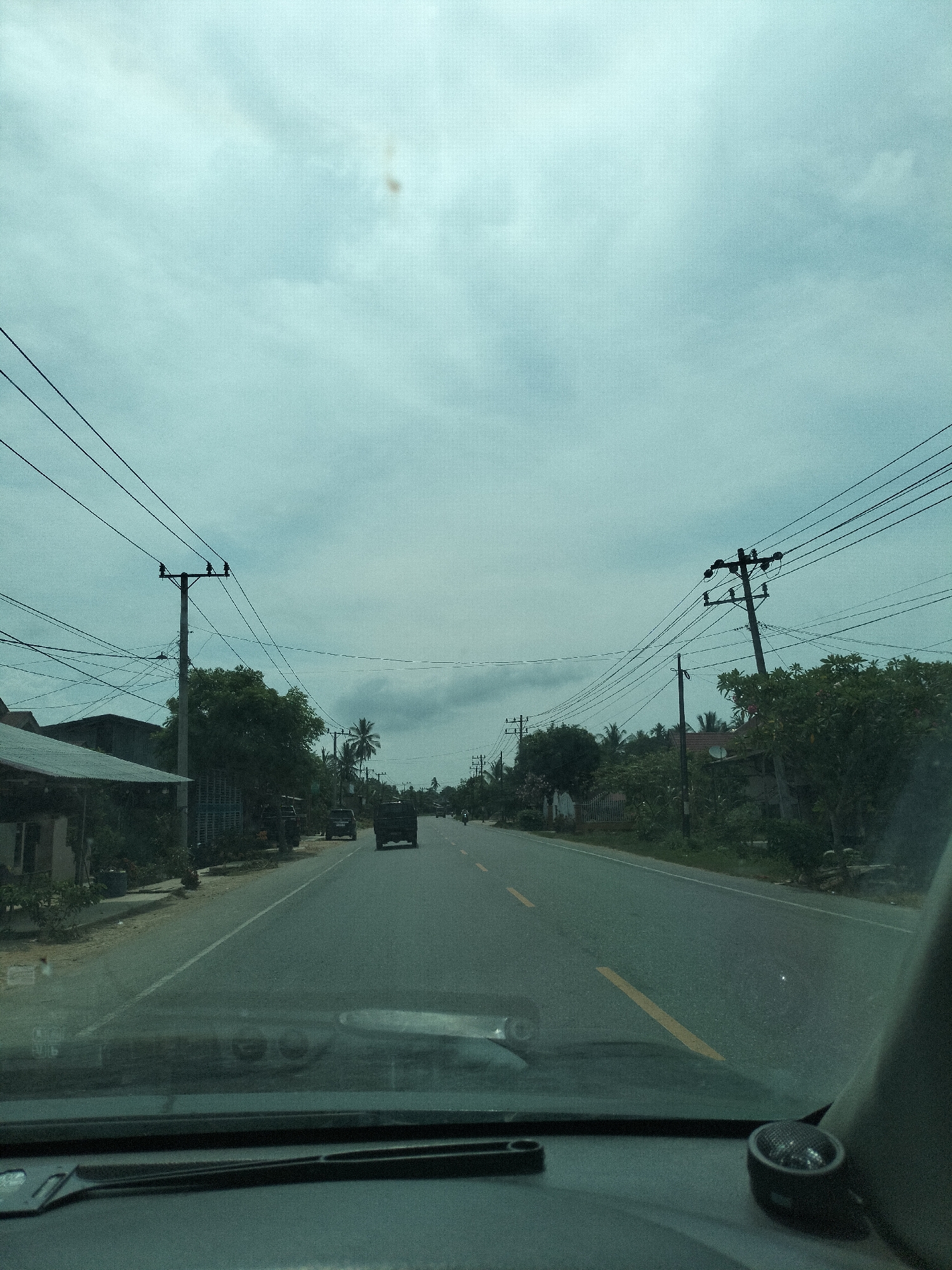 The image depicts a view from inside a vehicle traveling on a two-lane paved road, likely in Putoh Sa, Indonesia, given the context. The road is bordered by vegetation, including trees and palm trees, and sporadic buildings with corrugated metal roofs line the left side. Utility poles with numerous wires are visible on both sides of the road. Several vehicles are present on the road: two dark-colored SUVs are driving ahead, with a darker truck in the distance on the right lane, and possibly a motorcycle further down the road. The sky is overcast with a pale, greenish-blue hue, and a faint brown smudge is visible in the upper center of the sky. The interior of the vehicle is visible in the foreground, showing the dashboard and windshield.