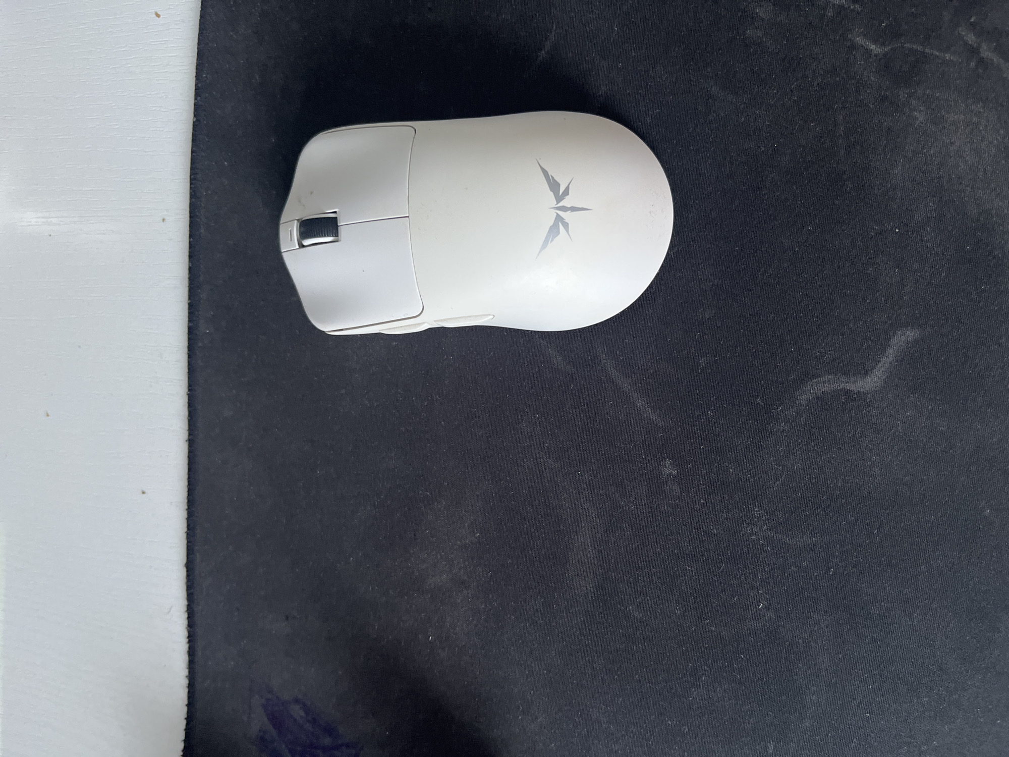 A white gaming mouse with a silver winged emblem is placed on a black mousepad. The mousepad is situated on a light-colored surface, possibly a desk or shelf. The background is out of focus, suggesting the photograph was taken indoors. There are no people present in the image. The location of this scene in Saratov, Russia is not provided.