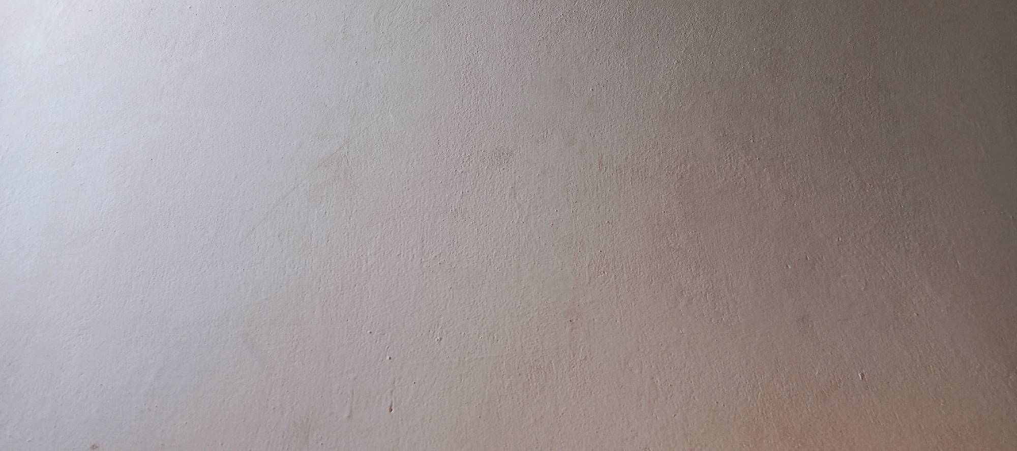 The image displays a close-up, flat-on view of a textured, light-colored wall, possibly painted plaster or concrete. The surface exhibits a slightly uneven texture with subtle variations in color and shade, suggesting a matte finish. There are no discernible objects, people, or actions visible within the frame. The lighting appears to be soft and diffuse, casting a gentle gradient of light across the wall, with the right side appearing slightly warmer or more illuminated than the left. The image is a generic representation of an interior or exterior wall surface and does not contain any elements that would allow for its localization to Miana Thub, Pakistan, or any other specific geographical location.