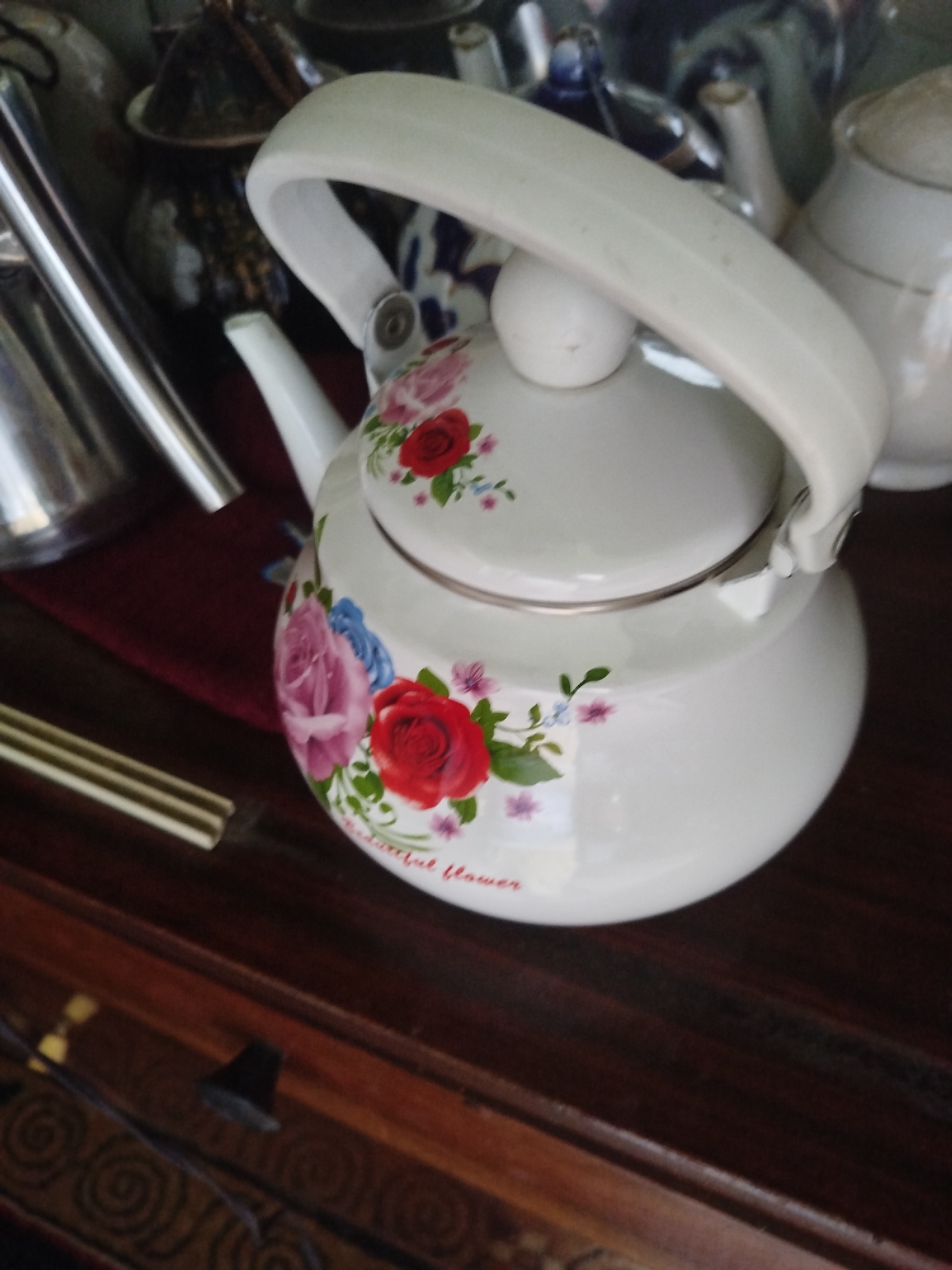 This is a close-up, eye-level shot of a white enamel teapot decorated with floral patterns. The teapot is positioned on a dark wooden surface, likely a table or shelf, which shows some wear and reflections. The teapot's handle is a prominent curved white piece, and it has a rounded lid. The floral design includes red and pink roses, along with smaller blue and pink flowers and green leaves. Some text, partially obscured, appears to be written in red on the lower part of the teapot, possibly "Beautiful Flower." In the background, out of focus, are other ceramic or metal objects, including what appears to be another teapot or a pitcher. The lighting is soft, suggesting an indoor setting. The location is identified as Sülüktü City, Kyrgyzstan.
