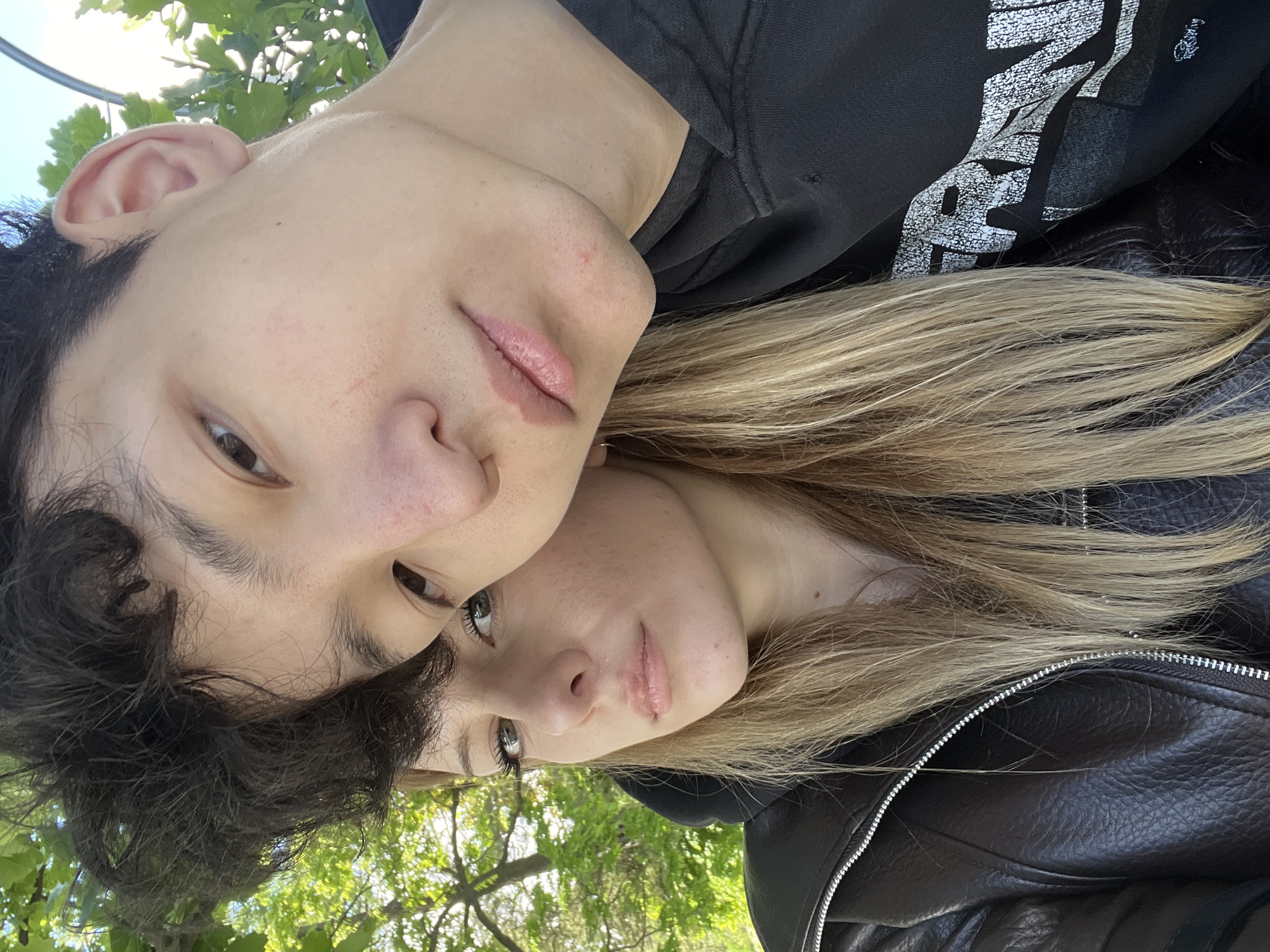 A close-up, low-angle shot depicts two individuals in an outdoor setting. The person on the left has dark hair and is wearing a black hoodie with white text printed on it. Their face is oriented towards the camera. The person on the right has blonde hair and is wearing a black leather jacket. Their face is also visible. The background reveals green leaves and sky, suggesting a location underneath a tree. The city and other location details are not known.