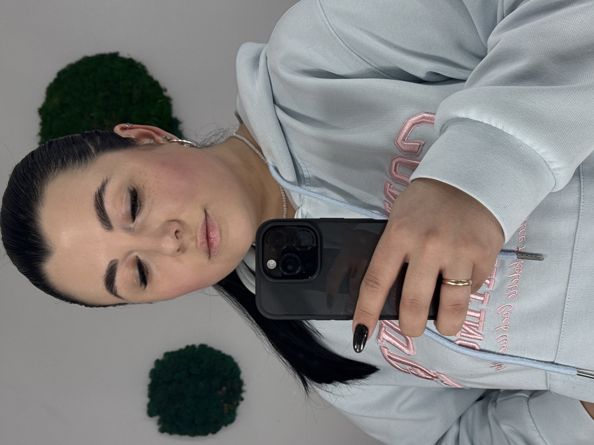 A woman is taking a selfie indoors. She has dark hair pulled back, fair skin, and is wearing a light blue hooded sweatshirt with pink lettering. She is holding a black smartphone in her right hand, which has a gold ring on her ring finger and a dark manicure. Behind her are two round green decorations mounted on the wall. The image does not offer sufficient information to infer the location of the photo being in Chaikovsky, Russia.