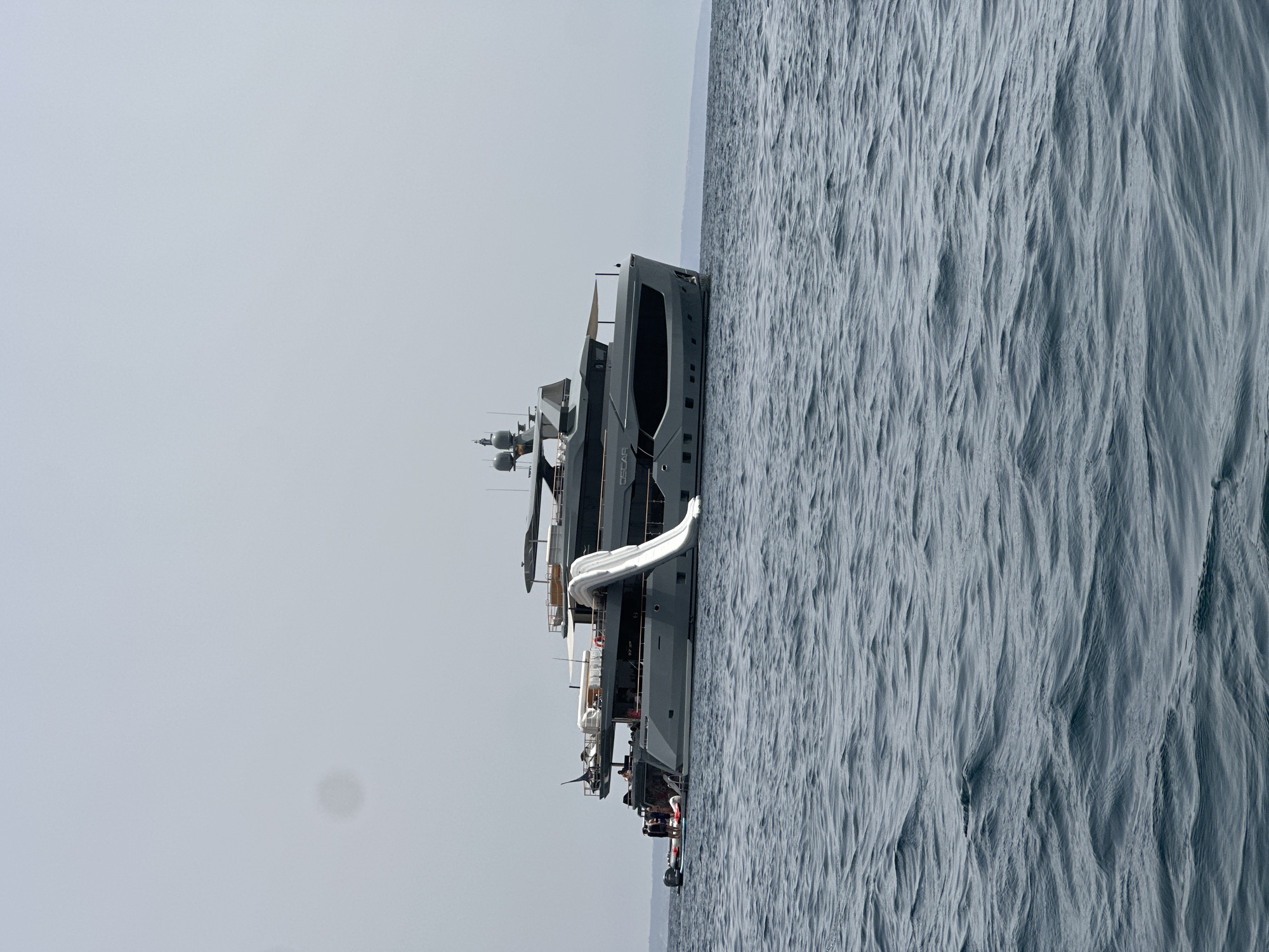 A large, grey motor yacht named "OSCAR" is anchored offshore in the waters of Calvià, Spain. The yacht has multiple decks, a prominent white waterslide extending from an upper deck to the water, and several inflatable watercraft are tied to its stern. A small group of people are visible on the aft deck of the yacht and near the watercraft. The sky is overcast, and the sea is calm with gentle ripples. The background shows a blurred outline of distant land.