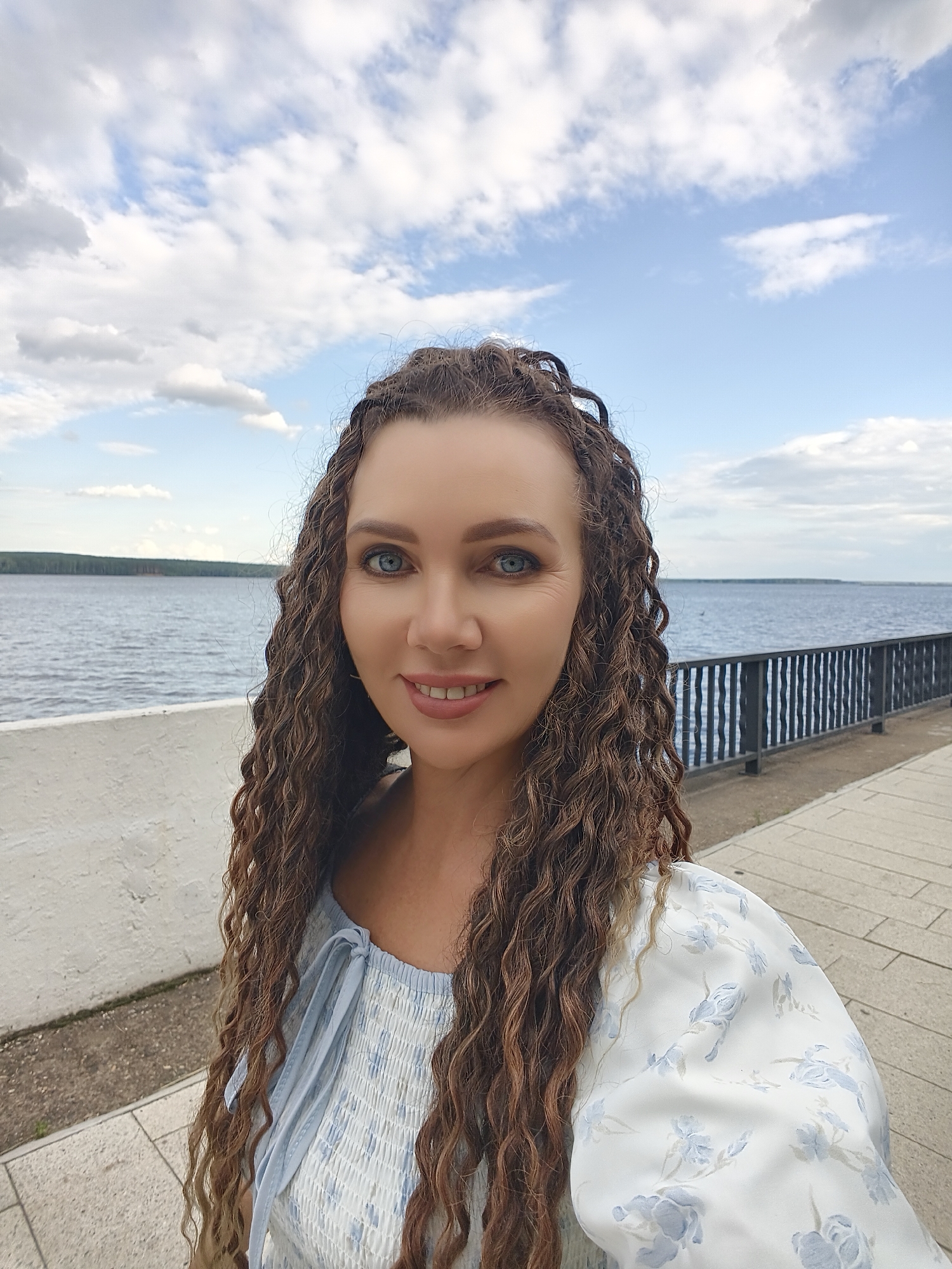A woman with long, curly brown hair, bright blue eyes, and a smiling expression takes a selfie outdoors. She is wearing a light blue and white floral print dress. The background shows a body of water, possibly a river, with a tree line in the distance. A paved walkway and a decorative metal railing are visible to the right of the woman, and a low, white wall is to her left. The sky above is partly cloudy. The scene appears to be a waterfront promenade in the city of Chaikovsky, Russia.