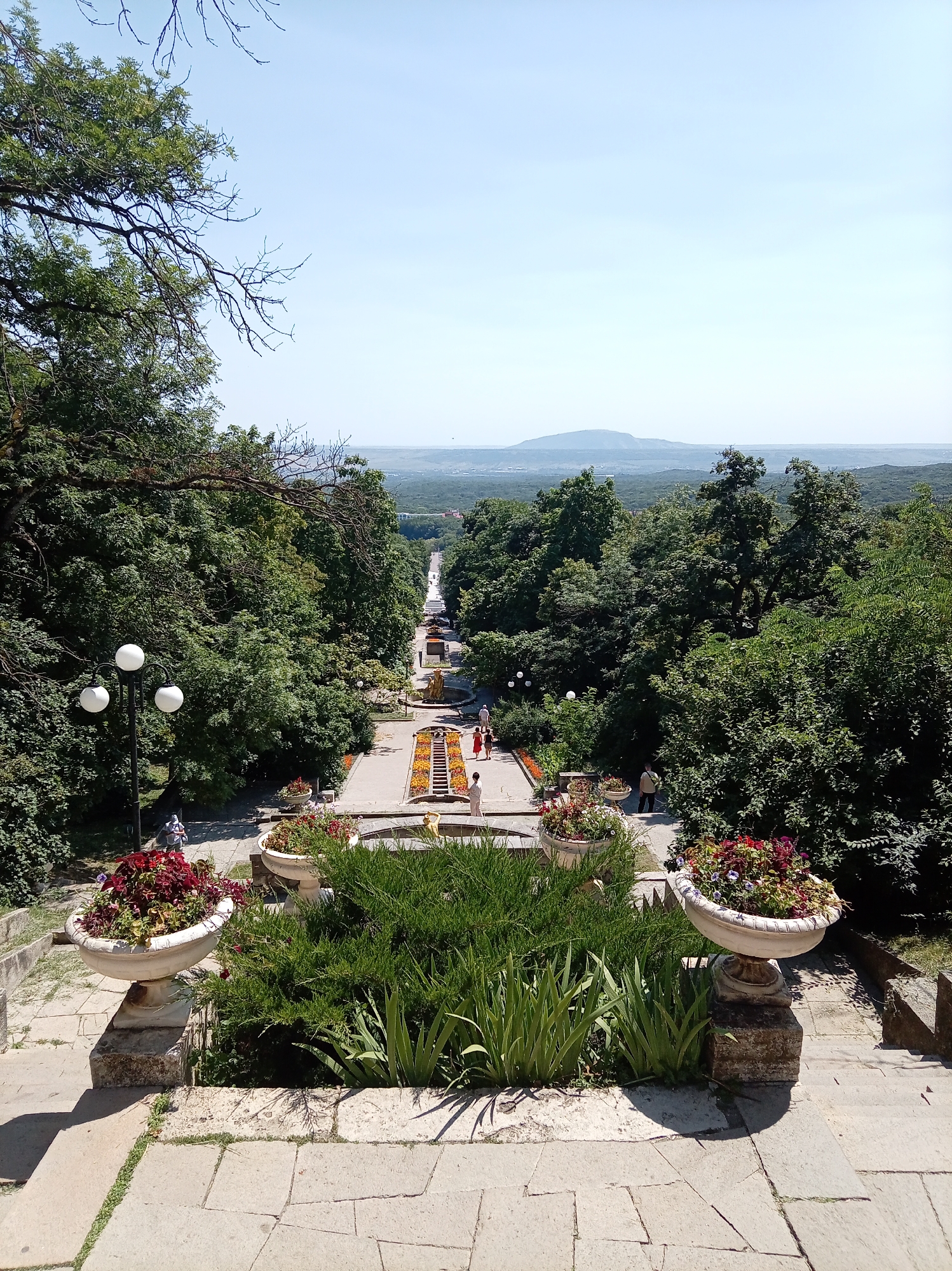 This is a high-angle, eye-level, medium shot of a long, grand staircase adorned with planters and leading down to a park-like setting, with a distant mountainous landscape under a clear sky. The staircase is made of light-colored stone, with wide steps and stone railings. Large, ornate planters filled with colorful flowers and greenery are strategically placed along the sides of the stairs and at intervals in the park area below. Several people are visible on the stairs and in the park, some walking and others seemingly resting.
To the left of the staircase, a vintage-style lamppost with three spherical lamps stands prominently. The surrounding area is densely wooded with lush green trees, creating a natural canopy. The park below features flower beds, pathways, and decorative elements, including a fountain or water feature with golden statues.
The distant background reveals a rolling landscape covered in trees, with a distinct, rounded mountain range in the far distance. The sky is a pale, cloudless blue, suggesting a clear and sunny day.
This scene is characteristic of the upper part of the cascade complex leading down to the main park in Zheleznovodsk, Russia, a well-known spa town in the Stavropol Krai region, famous for its mineral springs and scenic parks, often featuring similar ornate staircases and abundant greenery.