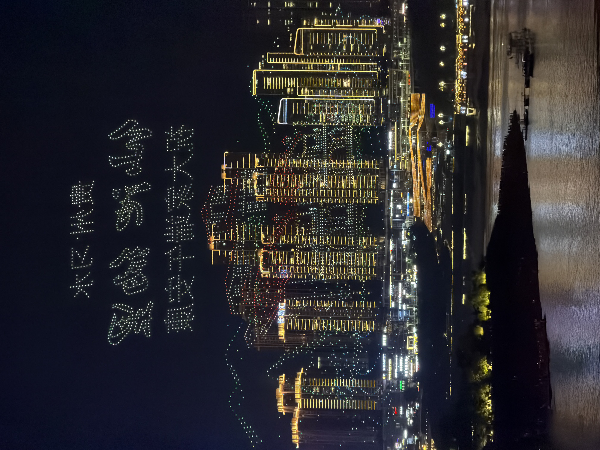 This is a nighttime aerial photograph taken in Yuzhong District, China. The main subjects are a drone light show and the surrounding cityscape and river. The drone light show displays Chinese characters formed by numerous illuminated drones against a dark sky. The characters appear to be a congratulatory message or a slogan.
Below the drone display, a densely built urban landscape is illuminated with many lights from buildings. The architecture suggests modern high-rise structures. A wide river flows beside the city, reflecting the lights. The time of day is clearly night, indicated by the darkness of the sky and the prominent artificial lighting. The weather appears calm, as the water surface of the river is relatively smooth.
Notable details include the precision of the drone formations, forming cohesive characters. The contrast between the dark sky and the bright lights of the city and drones creates a visually striking scene. The presence of the river and the extensive urban development suggests a significant metropolitan area.
The visible text consists of the Chinese characters formed by the drones. While specific characters cannot be definitively identified without further context or higher resolution, they are the primary textual element. The overall emotion evoked is one of celebration, technological advancement, and urban vibrancy.