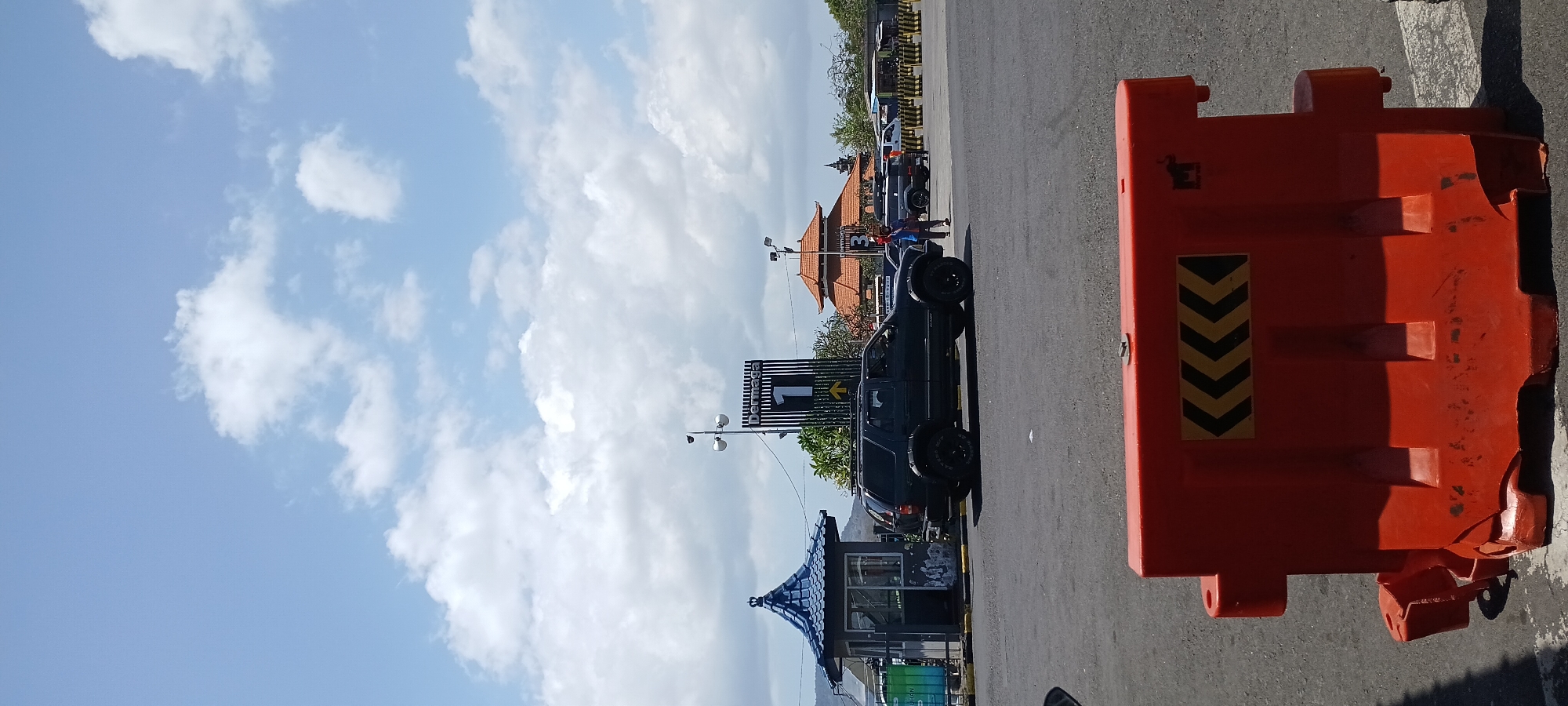 A dark-colored SUV is parked in an asphalt lot under a bright blue sky with scattered white clouds. The SUV is positioned near a small, blue, pagoda-style building with a decorative roof. In the background, a tall sign displaying "Dermaga 1" with an upward-pointing arrow is visible, along with buildings with traditional orange roofs and more vehicles. A bright orange traffic barrier with black and yellow chevron markings is in the foreground. Two individuals are standing near the SUV in the mid-ground. The scene appears to be a ferry terminal or port area in Melaya, Indonesia.
