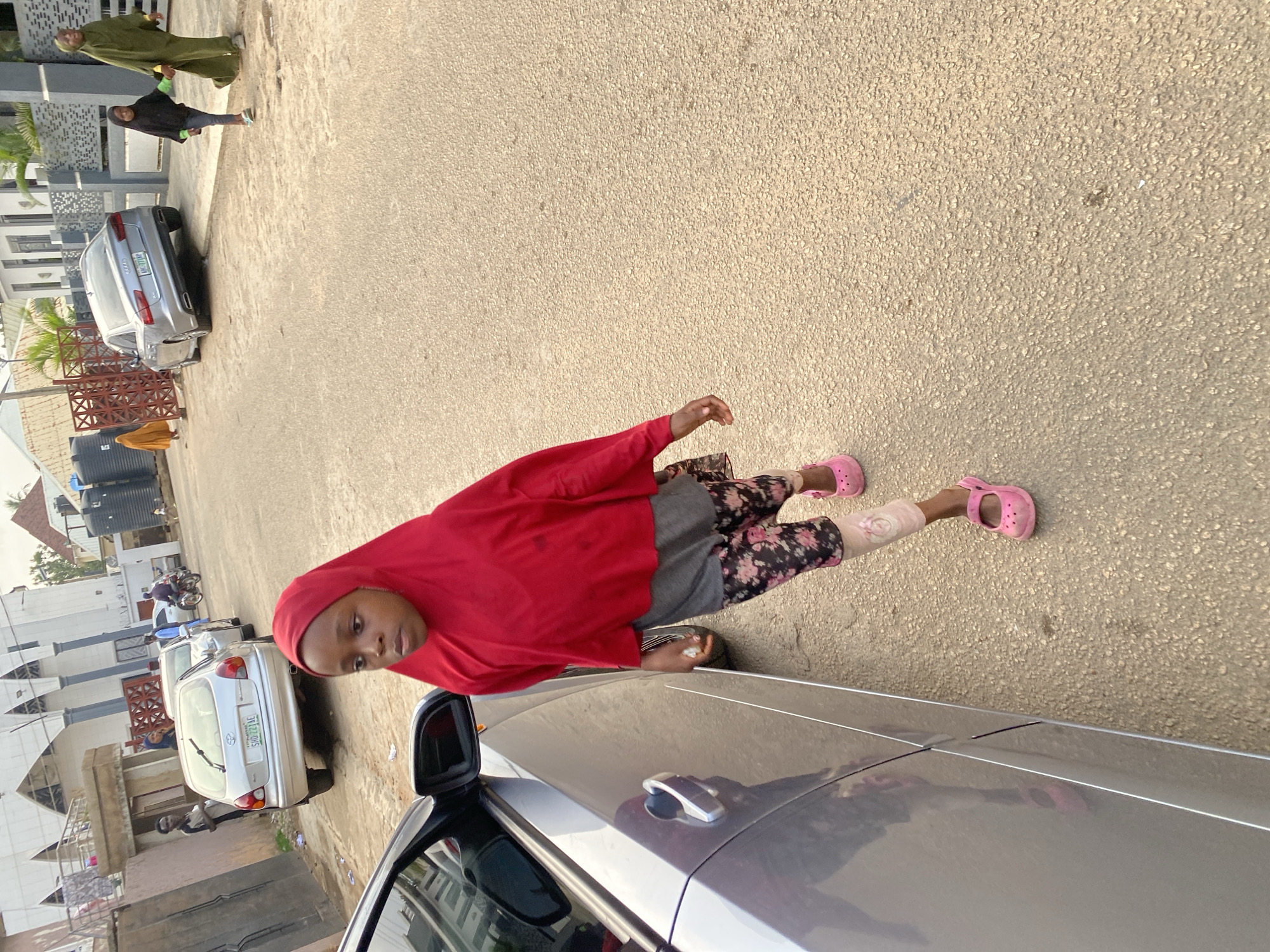 The image depicts a young girl wearing a red hijab and a floral-patterned skirt, walking on a paved road in Jos, Nigeria. She is accompanied by two women, one in a green hijab and another in a black hijab. A silver Audi car is parked on the right side of the road, while a silver Toyota is parked on the left. In the background, buildings and more cars are visible, suggesting an urban environment. The scene is illuminated by natural daylight.