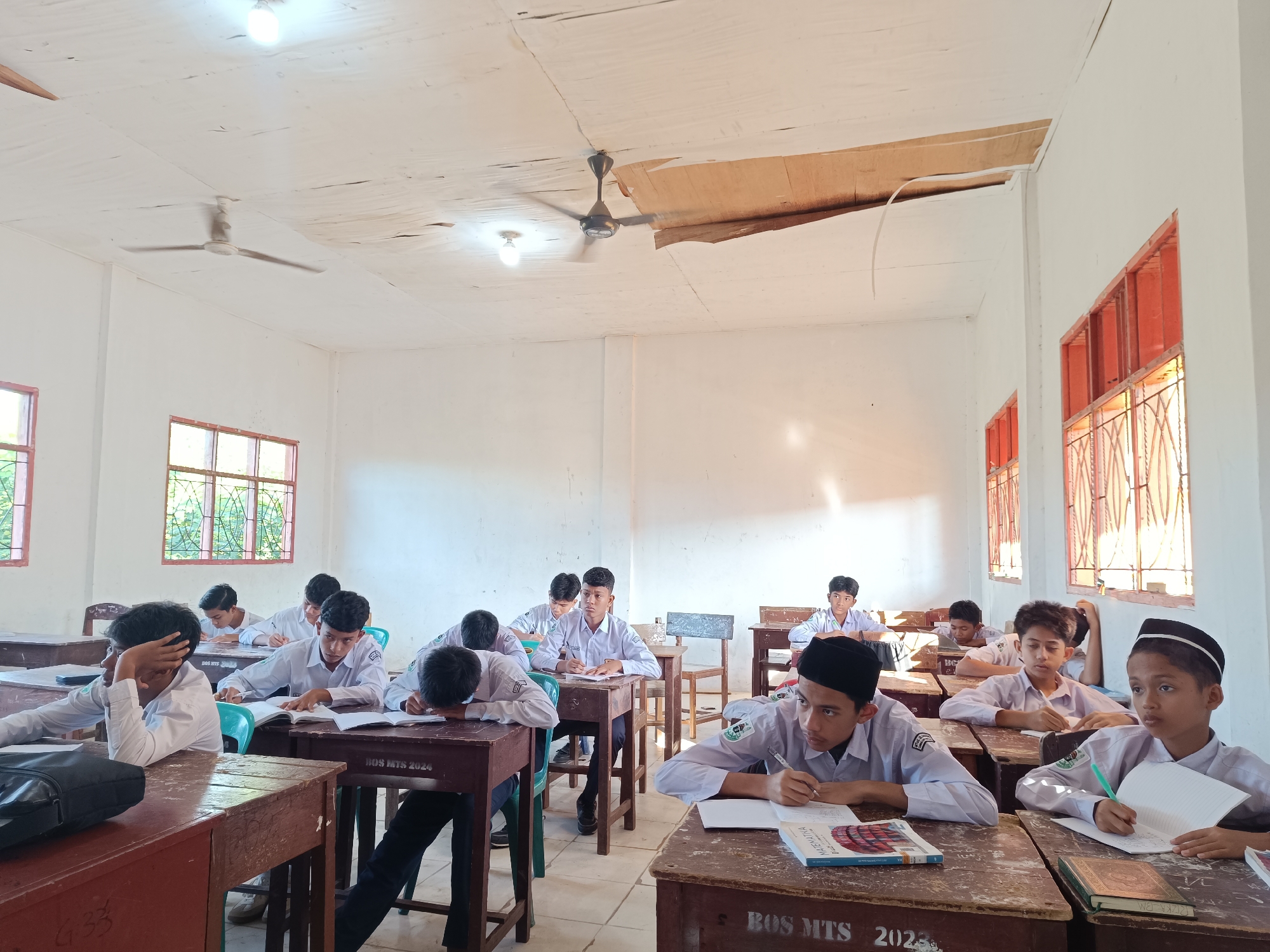 The image shows a classroom in Lhokseumawe, Indonesia. Around ten students are seated at wooden desks, wearing white shirts. Most are writing in notebooks or reading books, indicating they are engaged in academic activities. Some desks are labeled "BOS MTS 2024." The room is lit by ceiling lights and natural light from windows with metal bars. The ceiling appears damaged, with a section missing. There are ceiling fans in the room. The walls are painted white.