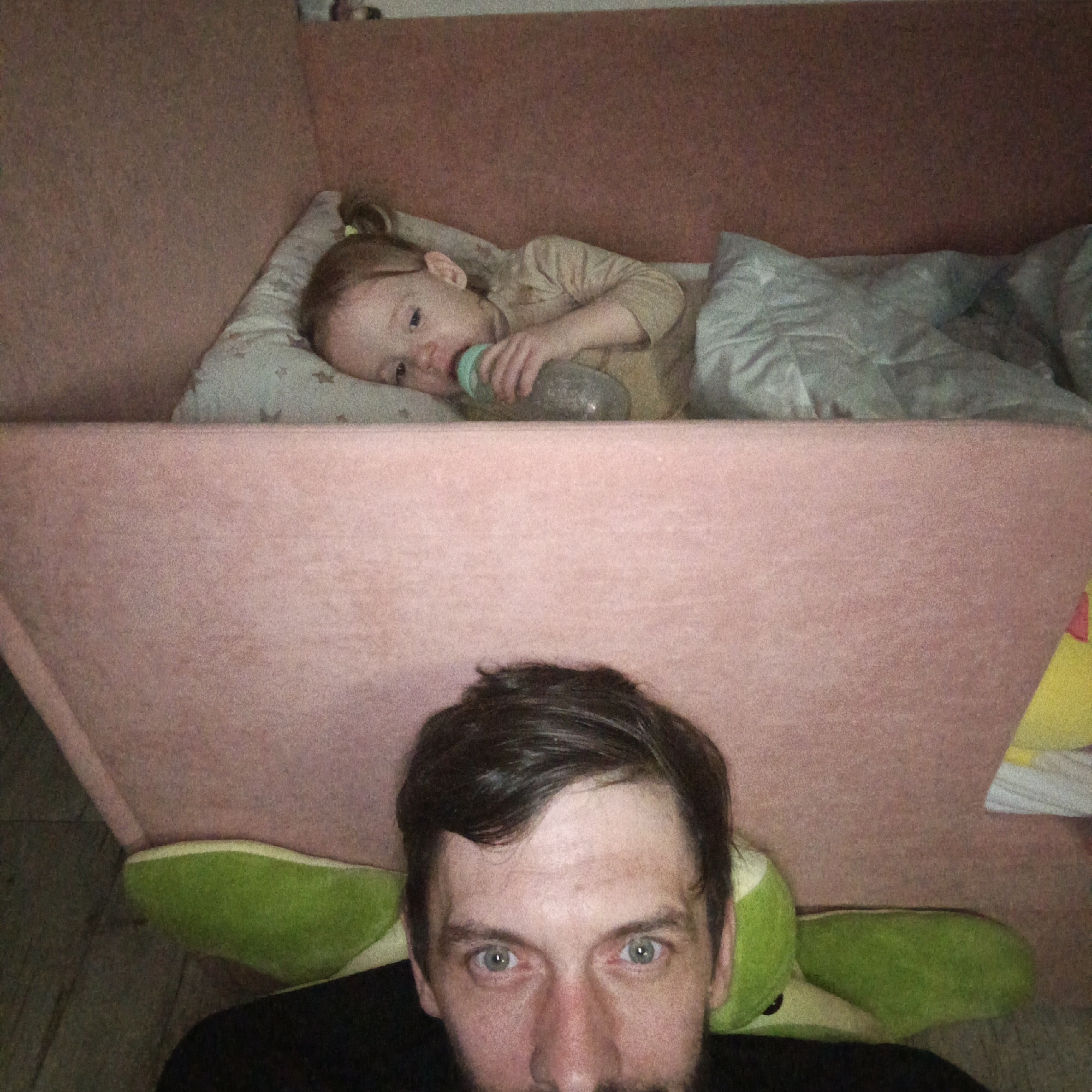 A photo shows a child in bed and an adult man looking at the camera. The child is lying on a pillow with a baby bottle in their mouth. A light-colored blanket covers part of their body. The bed has a raised, pink upholstered border. The man is positioned below and in front of the bed, with a green plush toy under his head. He is wearing a black shirt. The photo's angle suggests it was taken from a position slightly above the man and the head of the child's bed. The location is Марковский, Russia.