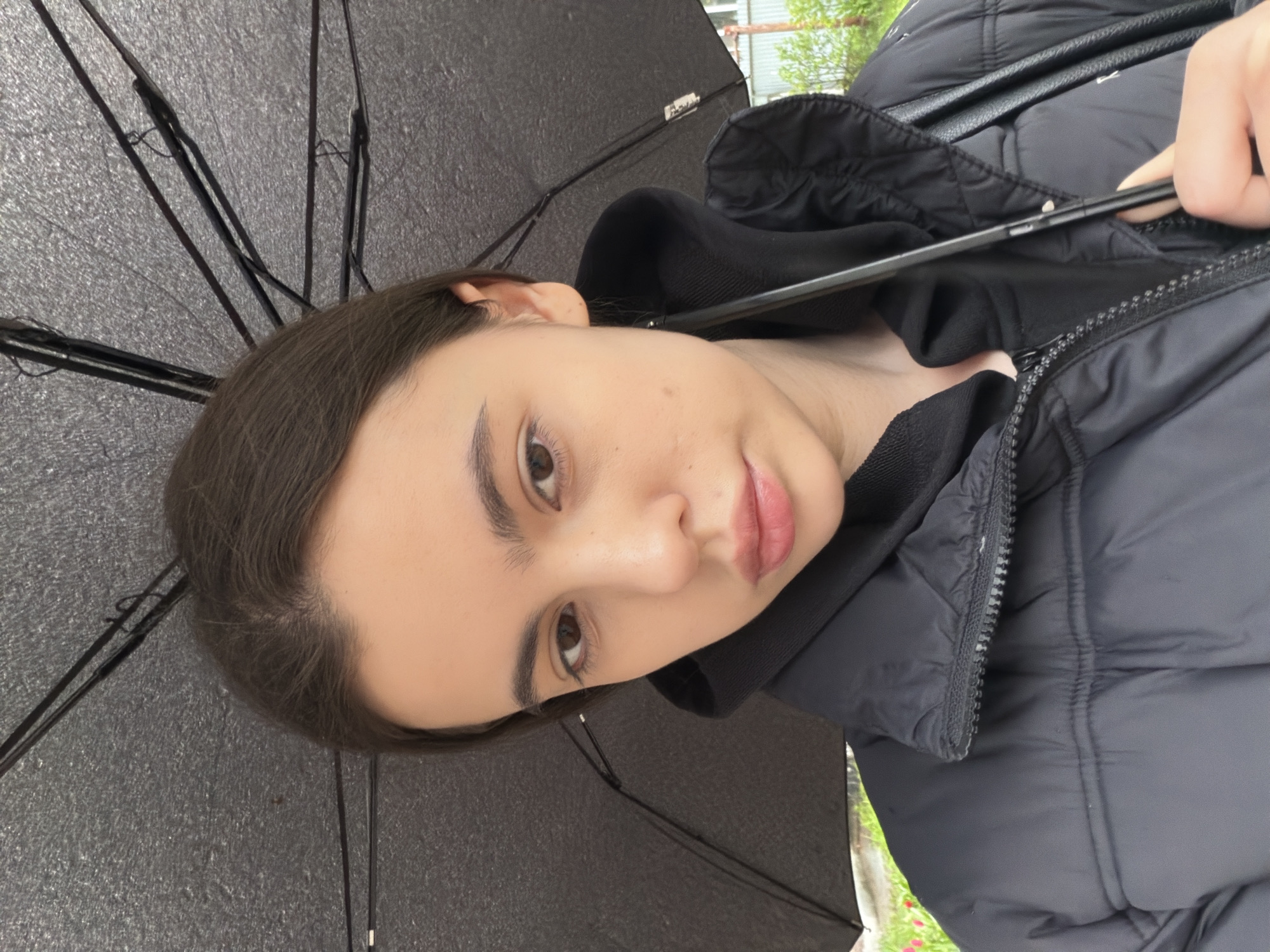 A young woman with brown hair is holding a black umbrella. She wears a black hooded sweatshirt under a puffy black jacket. The background shows blurry greenery and a building, possibly indicating an outdoor urban setting in Chaikovsky, Russia.