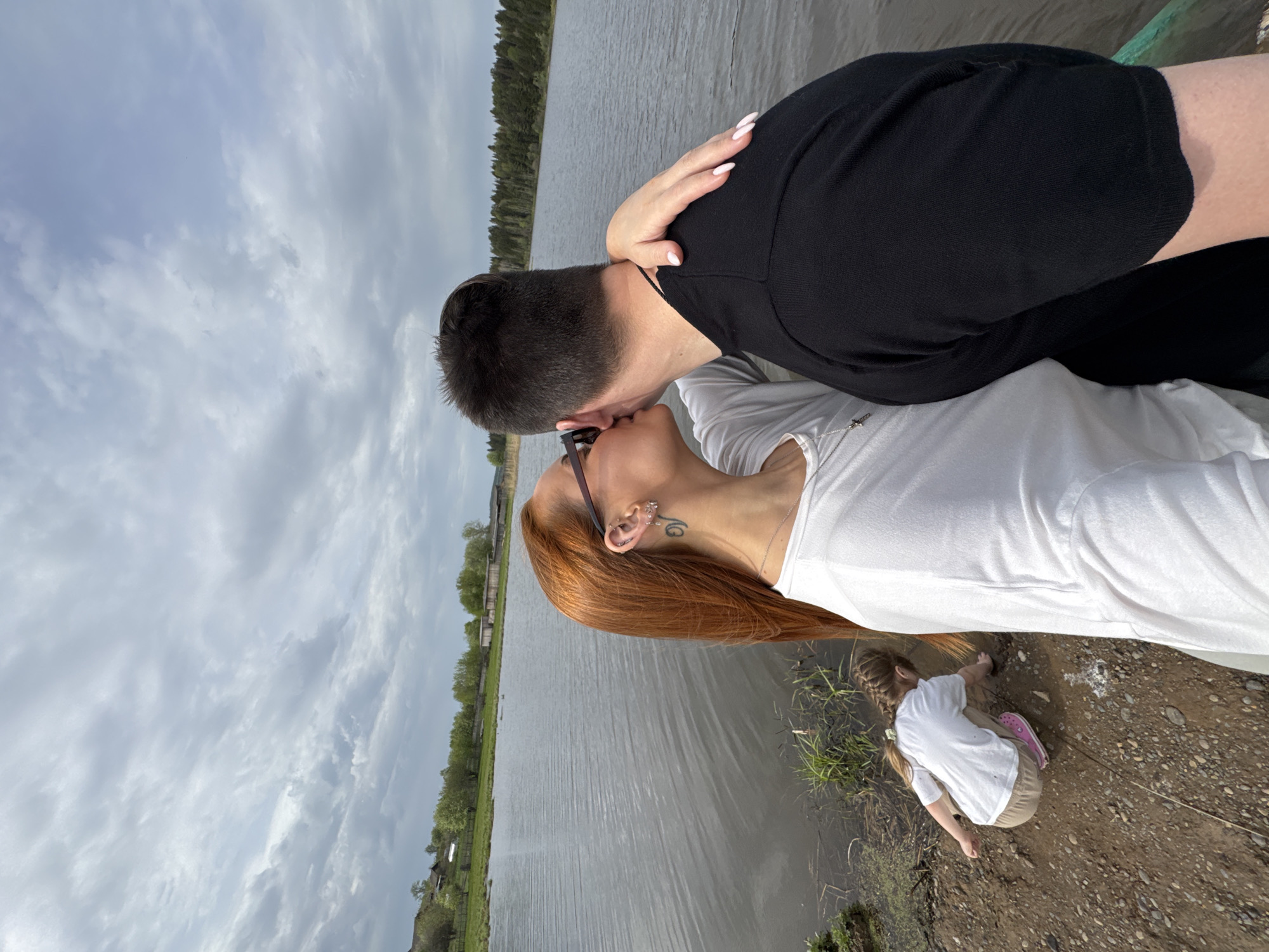 The image captures a couple kissing near a body of water in Nozhovka, Russia. The man wears a black shirt, while the woman has long, reddish-brown hair and a white shirt. A child in a white shirt and khaki pants kneels near the water's edge, seemingly examining the ground. The environment includes a lake, trees, and cloudy sky.