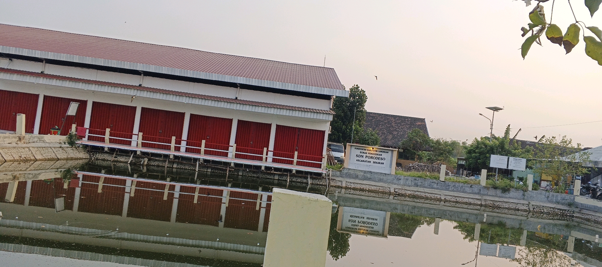 This is a photograph of the exterior of a school building, identified by a sign as "PEMERINTAH KABUPATEN LAMONGAN DINAS PENDIDIKAN SDN PORDOESO KECAMATAN SEKARAN". The building has a red roof and white walls with multiple red roll-up doors. It is situated alongside a body of water, possibly a pond or canal, which reflects the building and the sky. The water appears slightly murky and has some debris on its surface.
In the background, other buildings with dark tiled roofs and trees are visible. A street lamp with a solar panel is also present. The sky is overcast with a pale, warm hue, suggesting it is either early morning or late afternoon. A few birds are in flight. There are no people visible in the frame. The overall scene depicts a quiet, educational institution in a semi-rural or suburban setting. The location is explicitly stated as Porodeso, Sekaran District, Lamongan Regency, Indonesia.